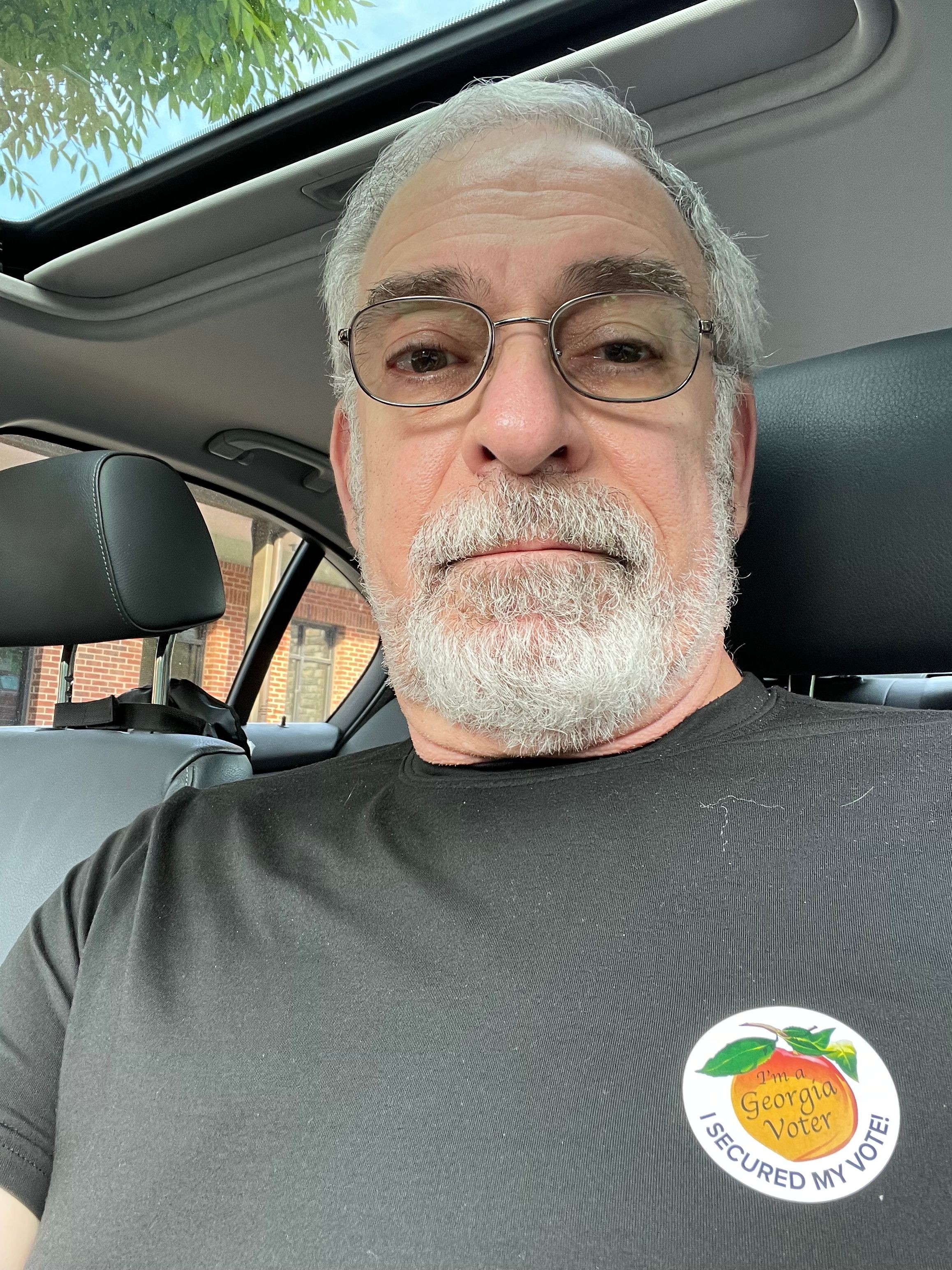 Older man with glasses and white beard sitting in a car, wearing a dark shirt with a sticker on it, looking at the camera.