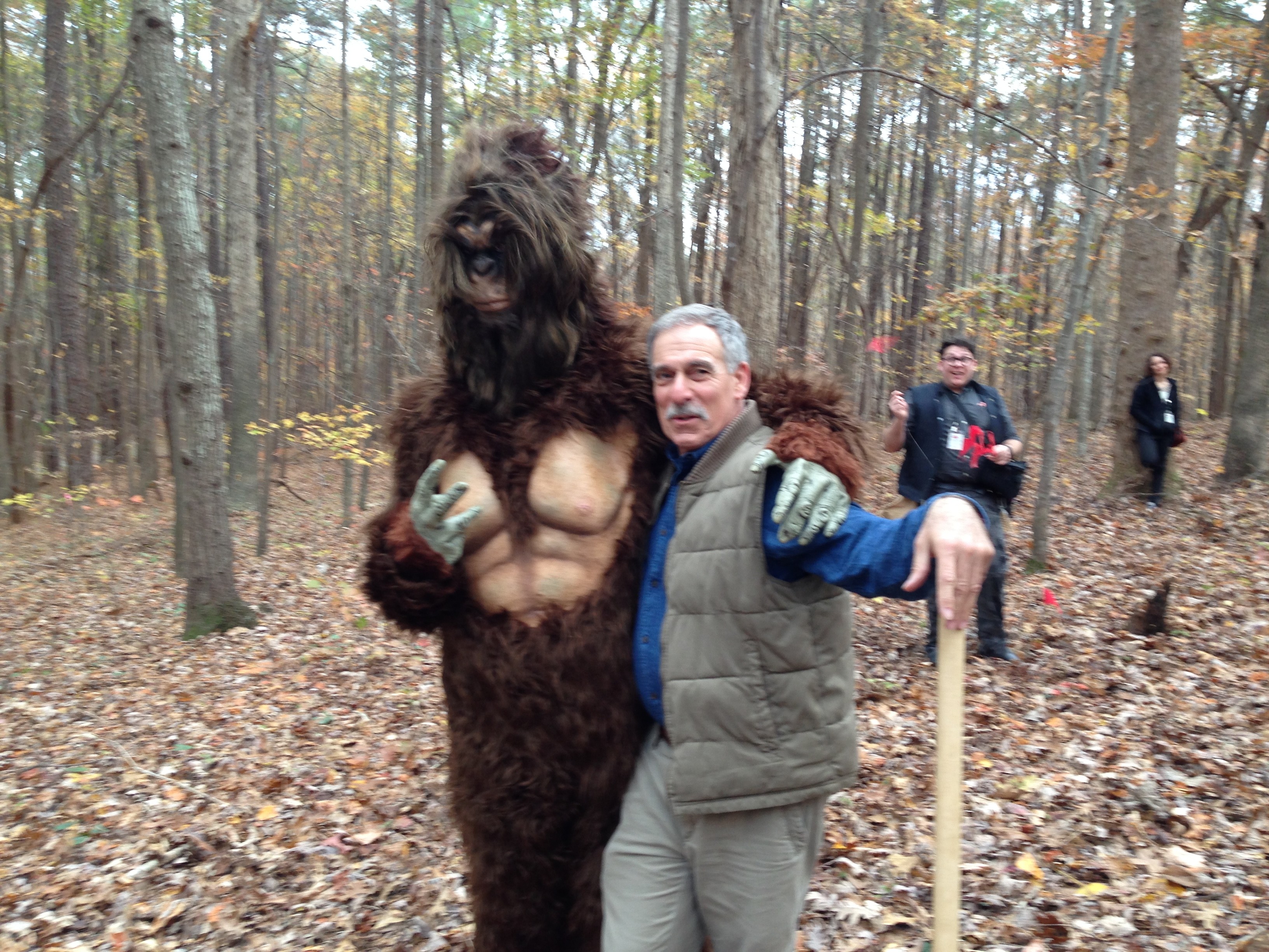 Person standing next to a person in a Bigfoot costume in a forest with fallen leaves.