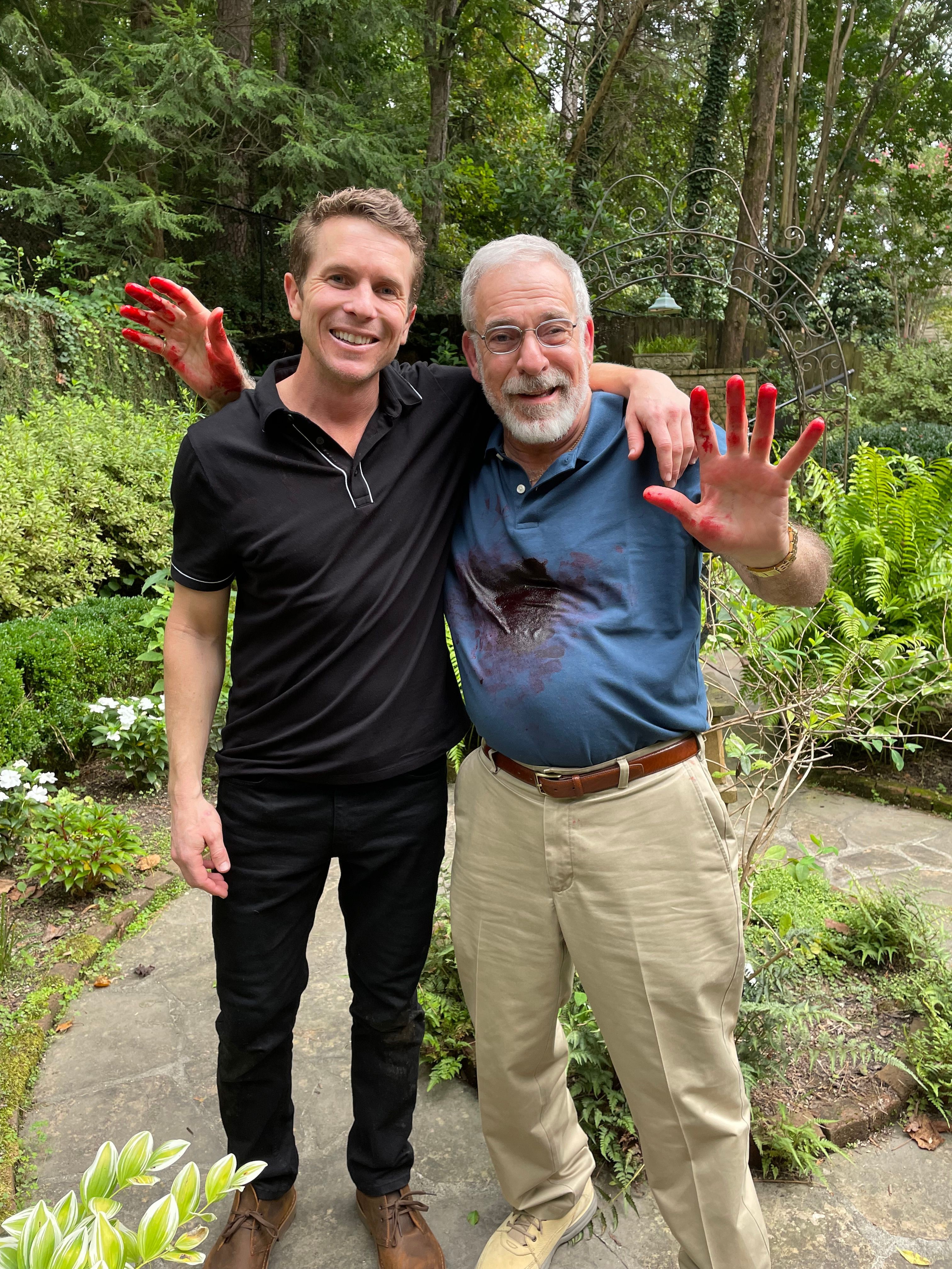 Two men standing outdoors in a garden, one older with gray hair and a beard, the other younger with short hair, both smiling and waving.