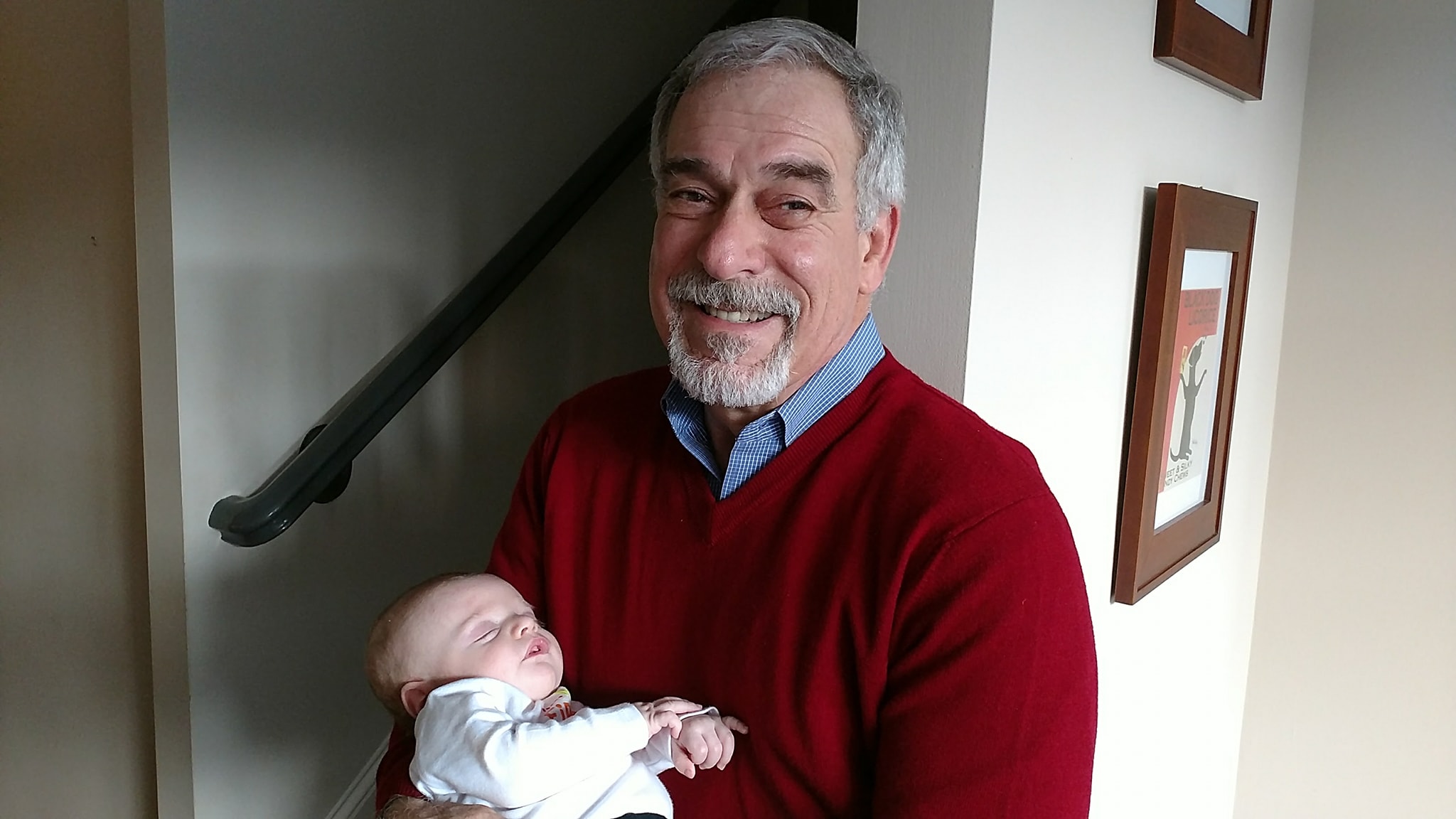 Older man holding a newborn baby indoors, smiling, with framed pictures on the wall behind him.