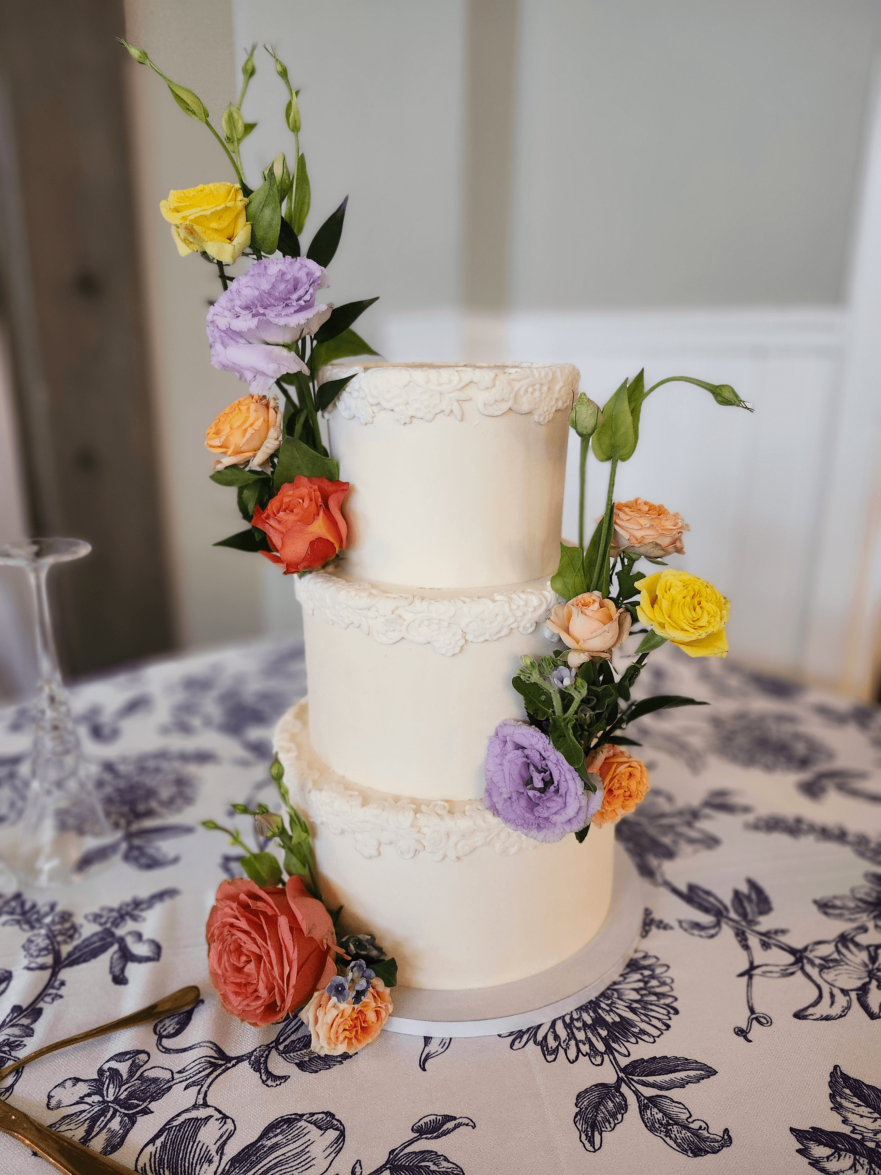 Three-tiered white wedding cake decorated with colorful flowers, on a table with a floral tablecloth.