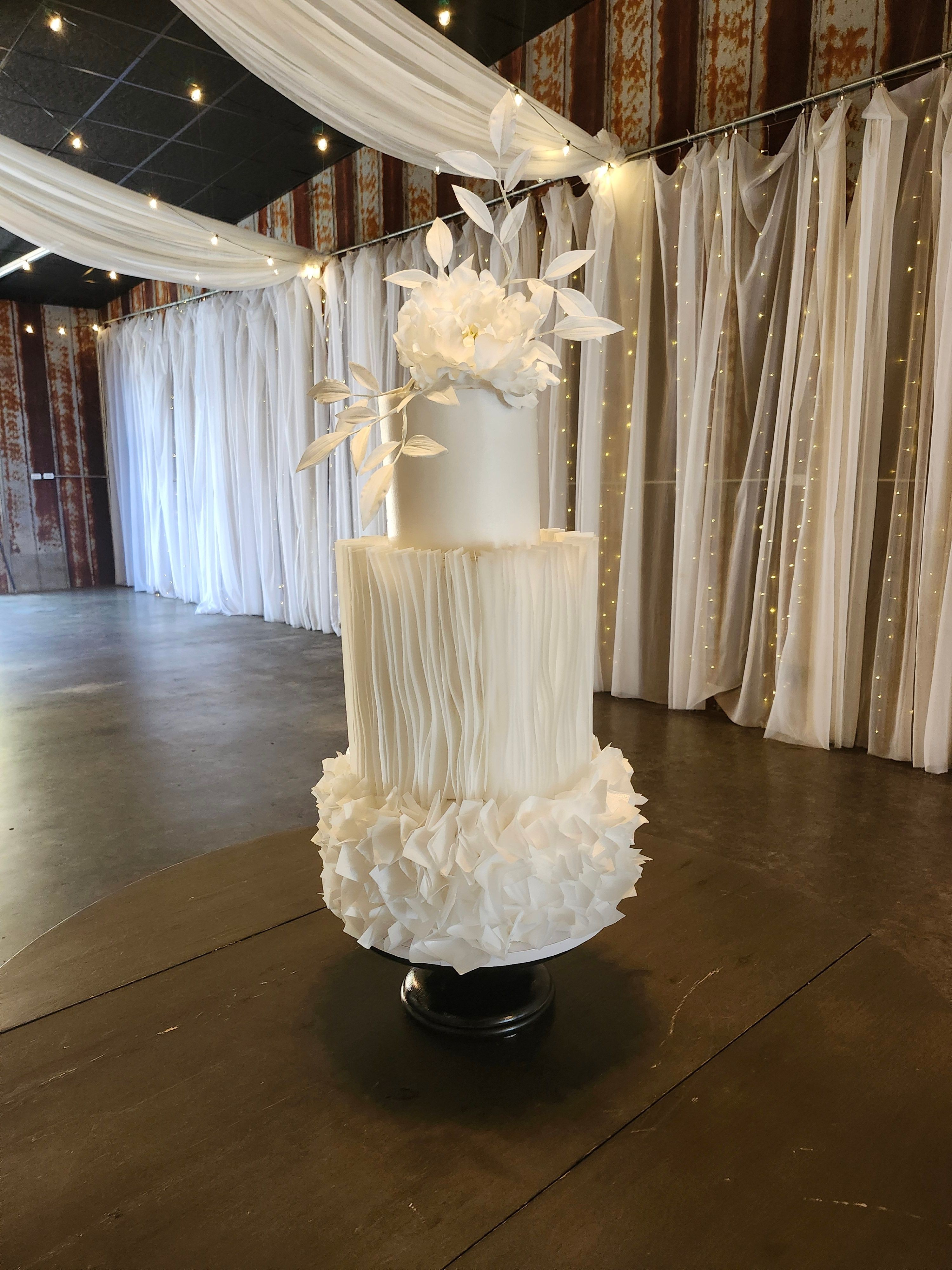 Tall white tiered wedding cake with floral decorations on top, placed on a dark wooden floor, with draped curtains in background.