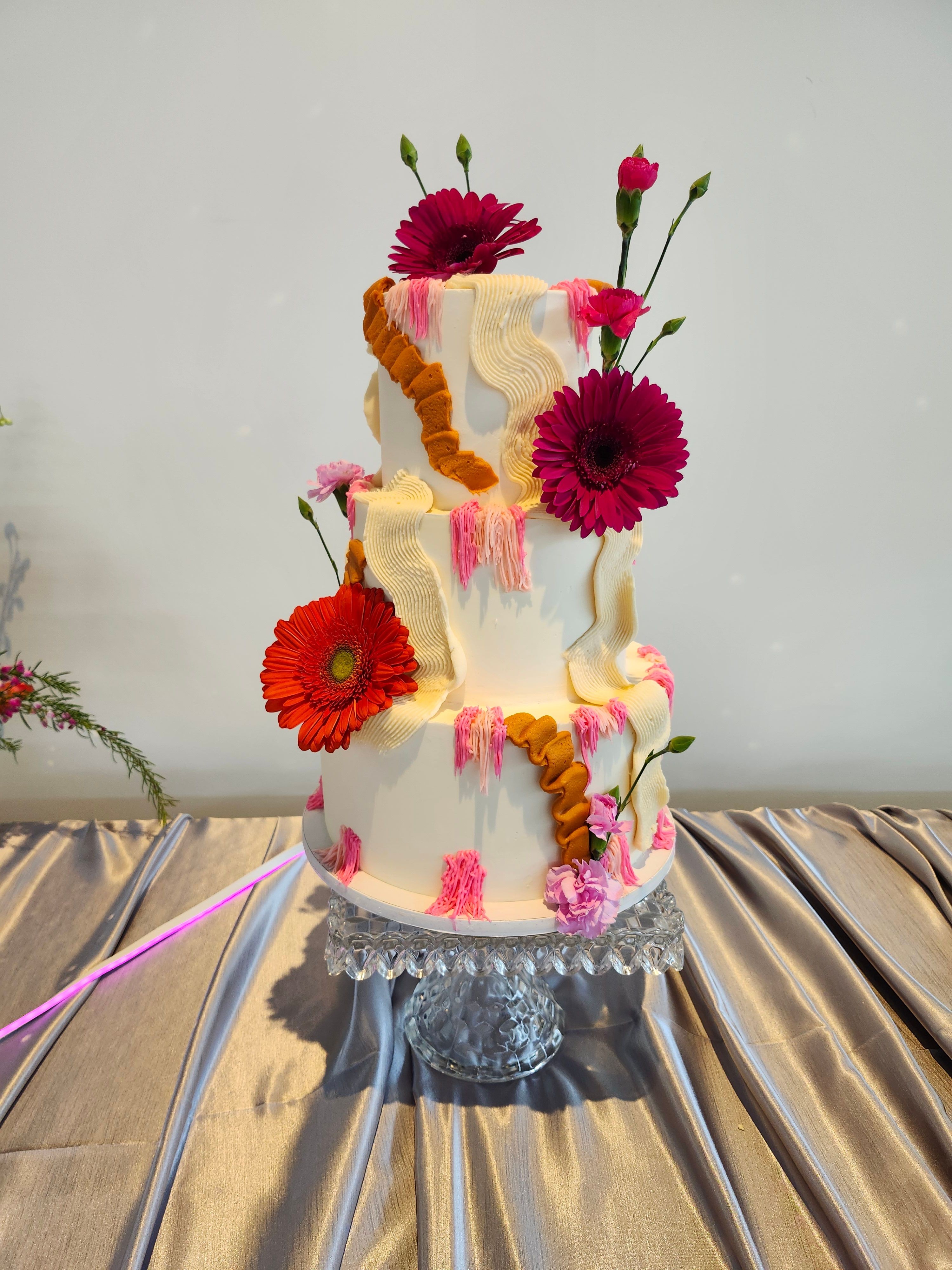 Cake decorated with pink and red flowers, placed on a table with a gold-colored draped cloth, with a white background.