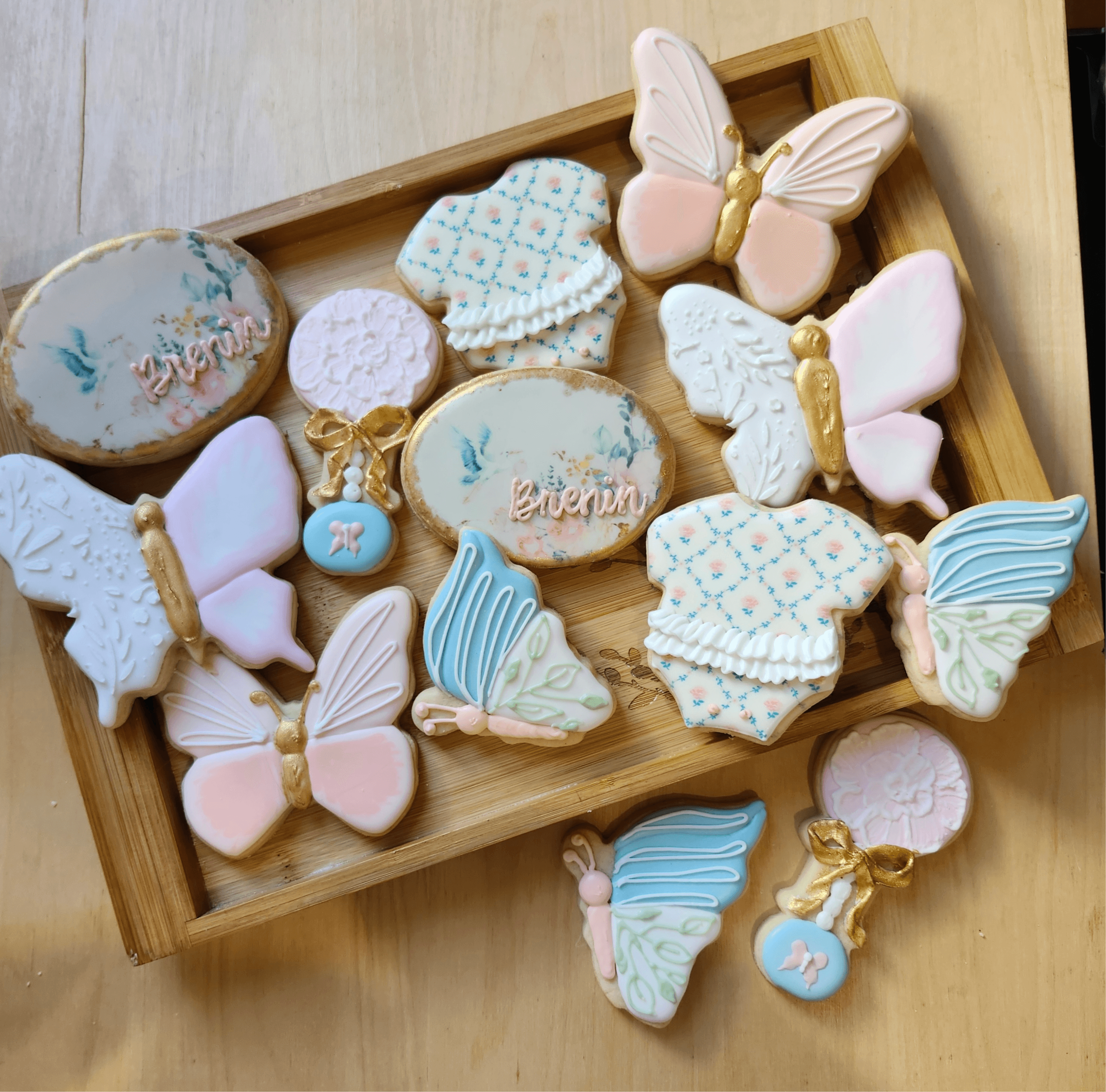 Decorated cookies with butterfly and cloud shapes, some with pastel colors and gold accents, arranged in a wooden tray.