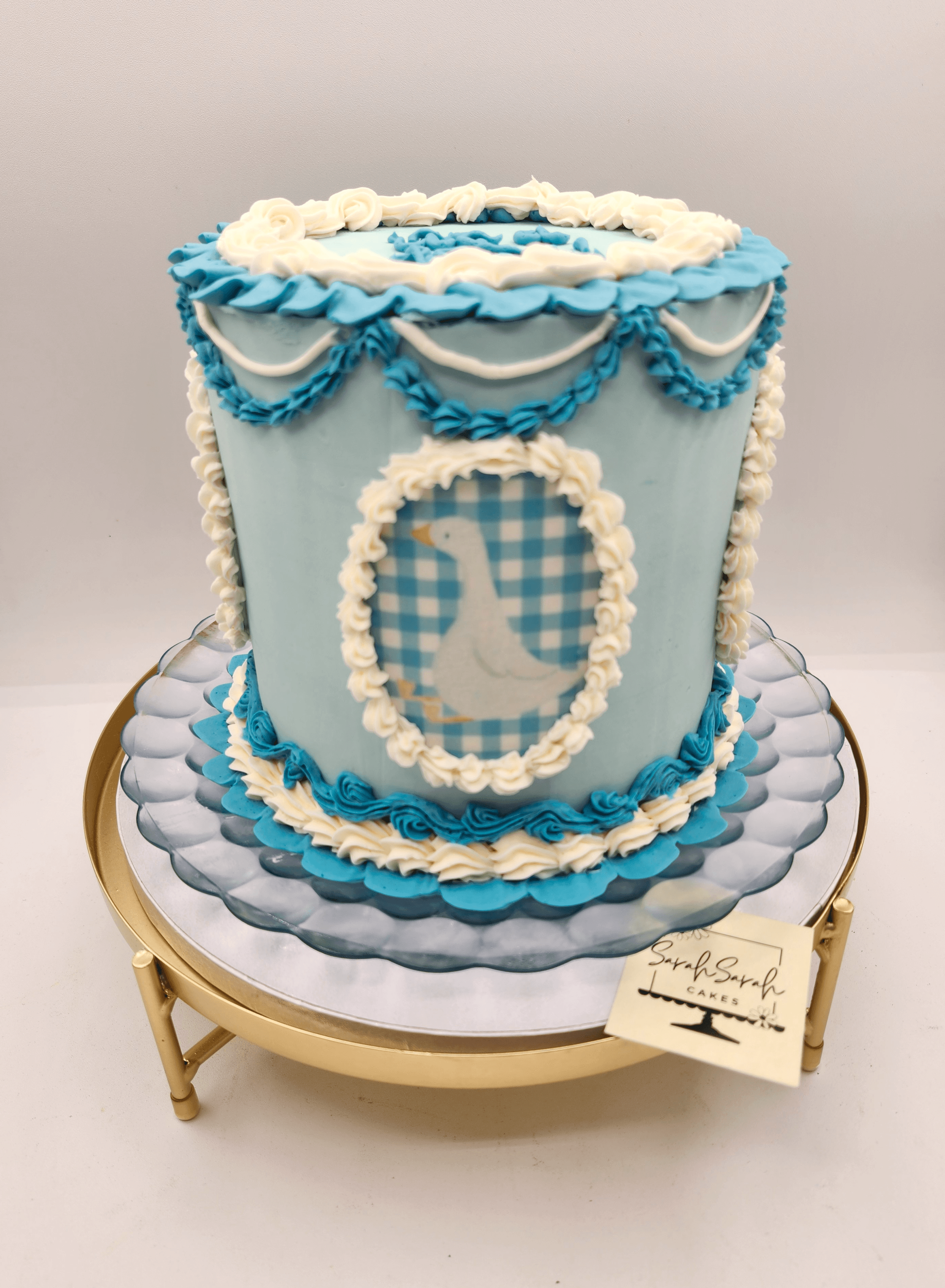 Decorative cake with blue and white icing, featuring a framed checkered pattern, on a round gold stand.