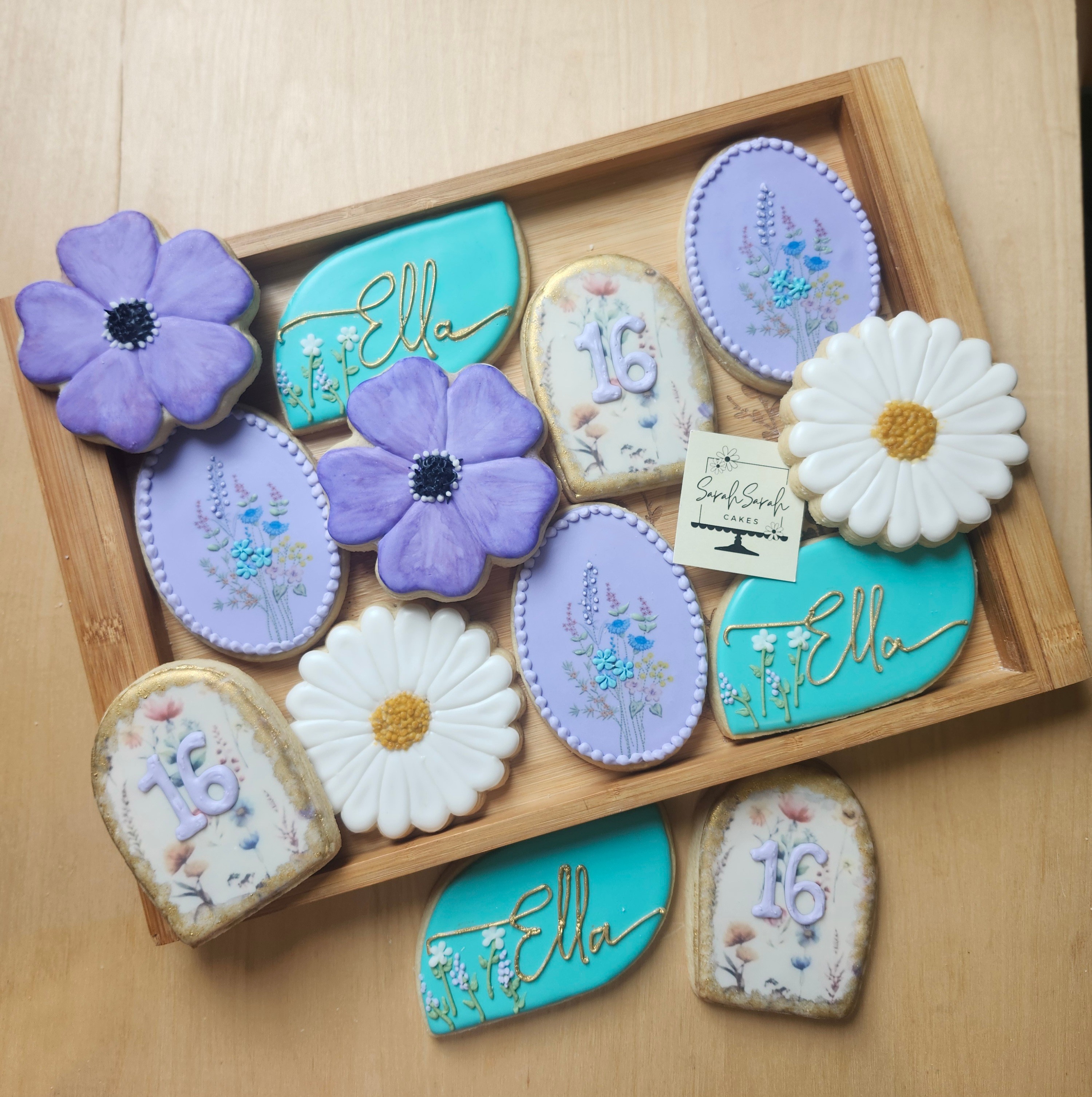 Wooden tray with decorated cookies and flowers, including purple, white, and blue cookies, on a light wooden surface.