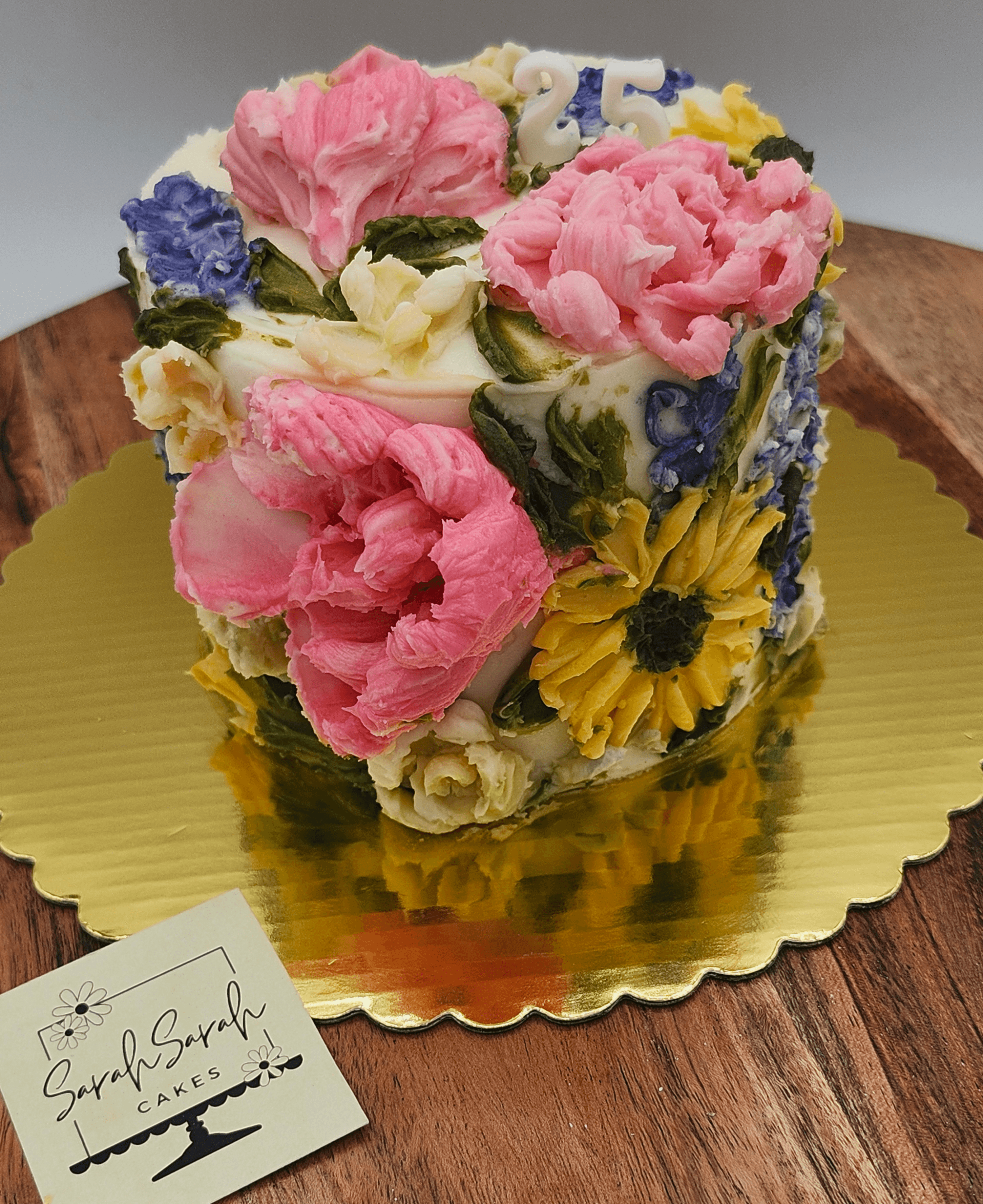 Decorative cake with pink, purple, yellow, and white flowers on a gold base, on a wooden surface.