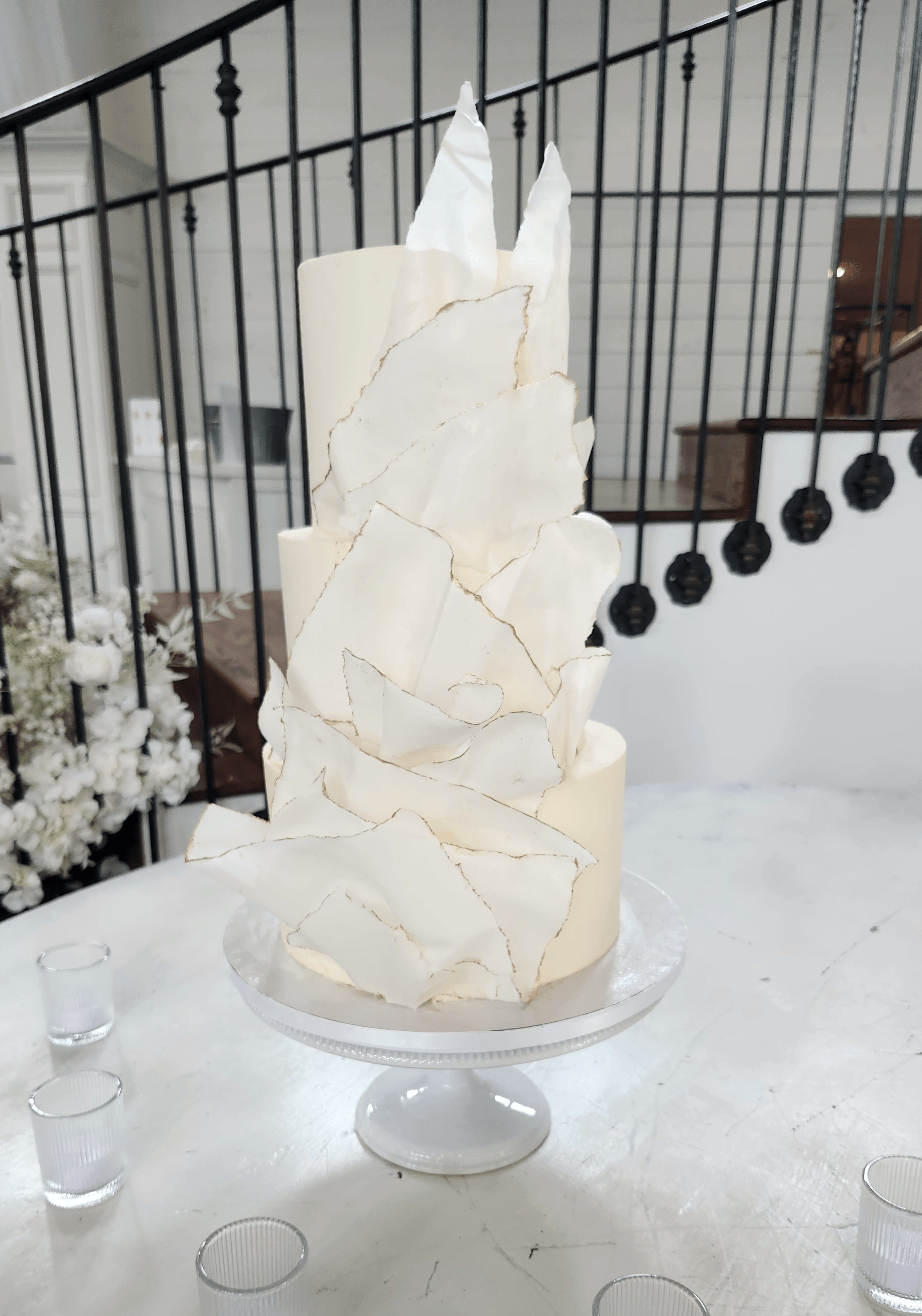 Tall white cake with irregular white decorations on a glass stand, set on a white surface with a black railing in background.