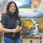 Woman holding paintbrushes standing in front of landscape painting in art studio.