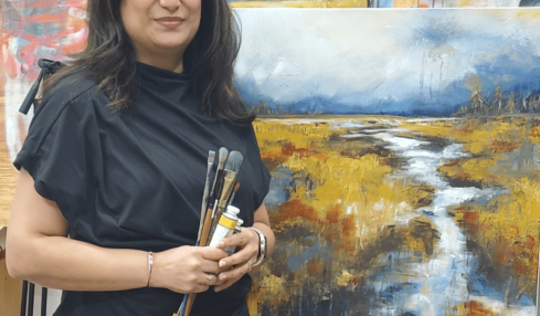 Woman holding paintbrushes standing in front of landscape painting in art studio.