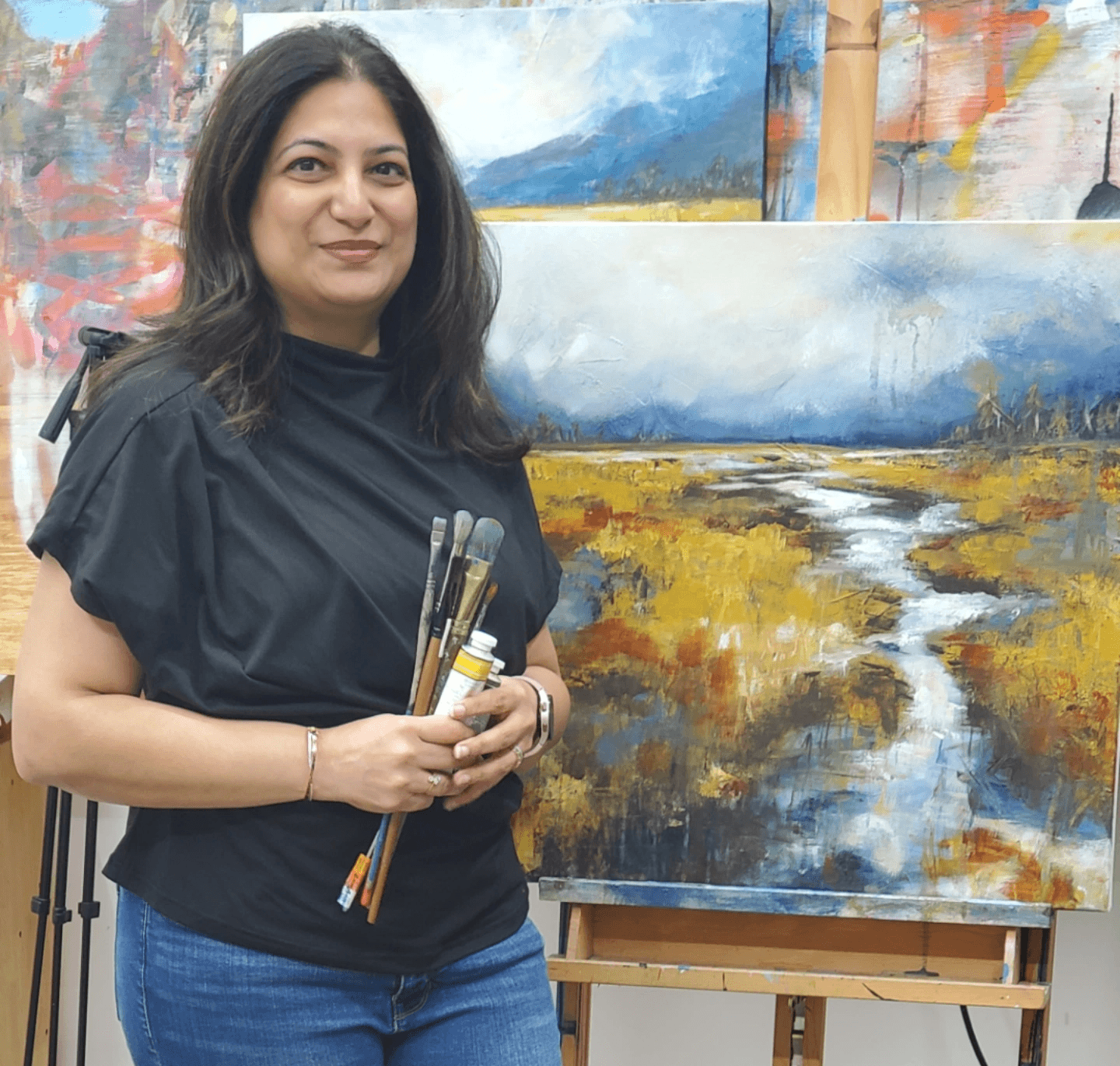 Woman holding paintbrushes standing in front of landscape painting in art studio.