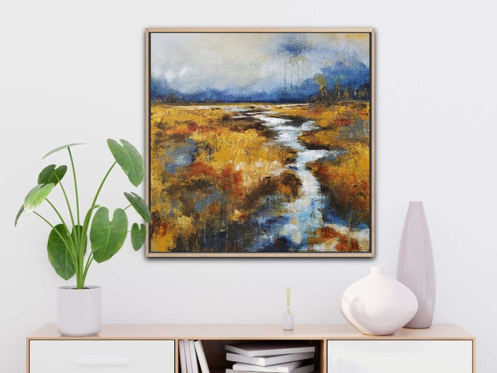 Landscape painting of a river flowing through a field with a cloudy sky, in a gold frame, on a white wall.