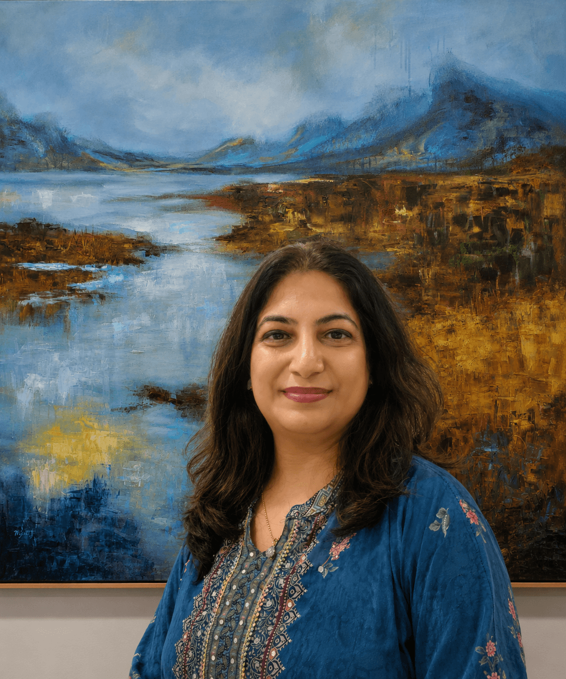 Woman with dark hair and a blue embroidered top standing in front of a landscape painting of mountains, water, and sky.