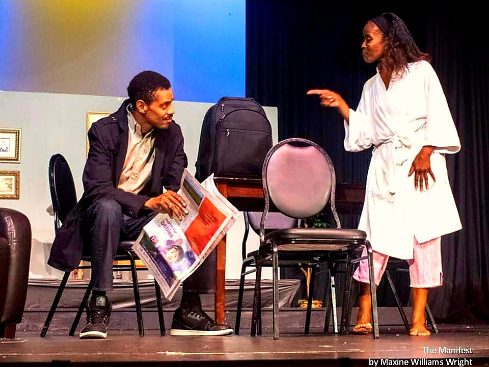 Two women engaged in conversation on stage, one seated holding a newspaper, the other standing gesturing, with chairs and a backpack nearby.