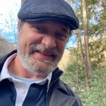Man outdoors wearing a flat cap, smiling, with trees and sky in background.