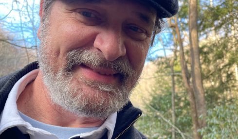 Man outdoors wearing a flat cap, smiling, with trees and sky in background.
