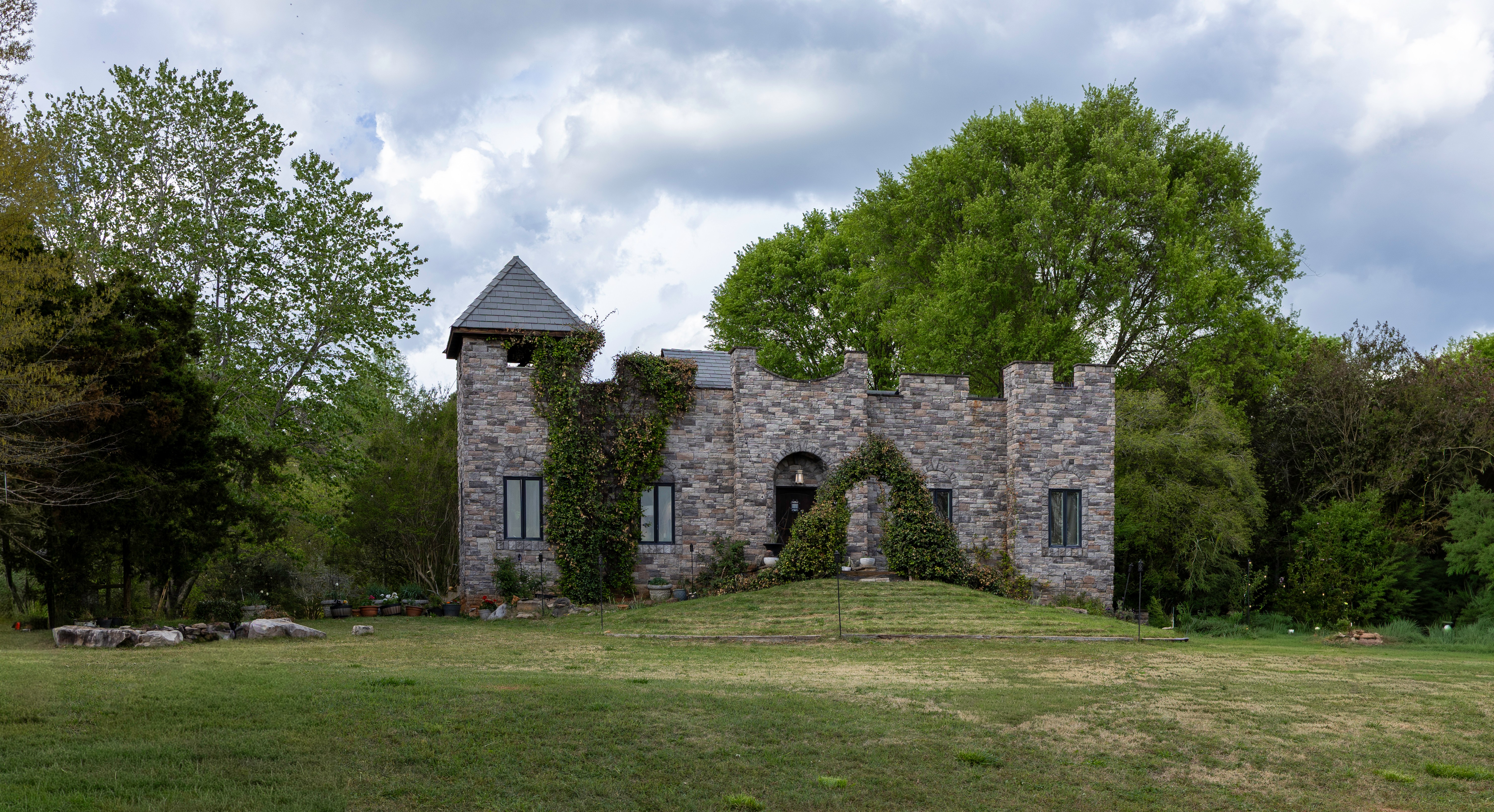 Stone castle with tower and arched entrance surrounded by trees and grass, under cloudy sky.