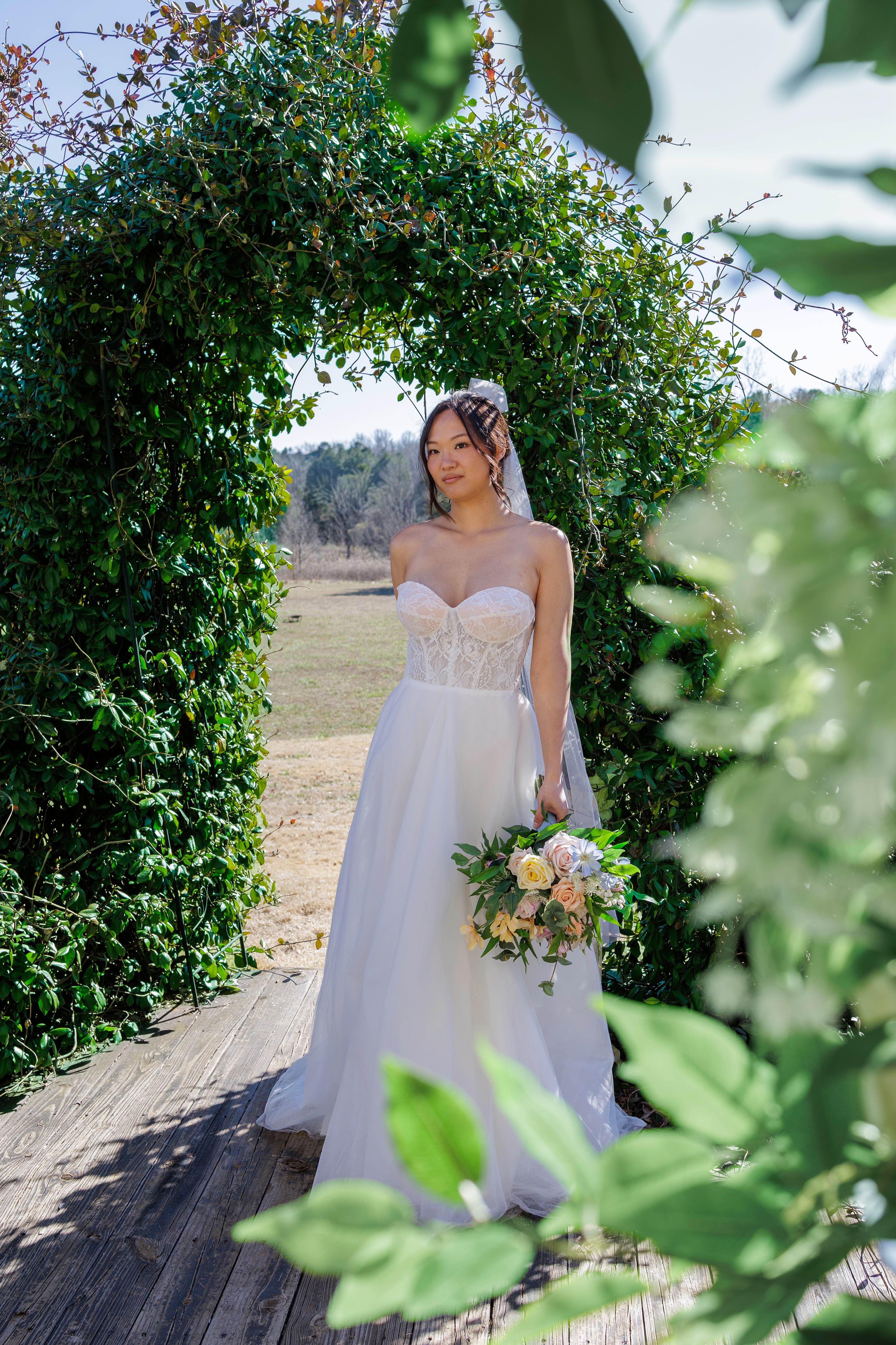 Bride in wedding dress holding bouquet stands outdoors under a green archway, sunlight on her face and dress.
