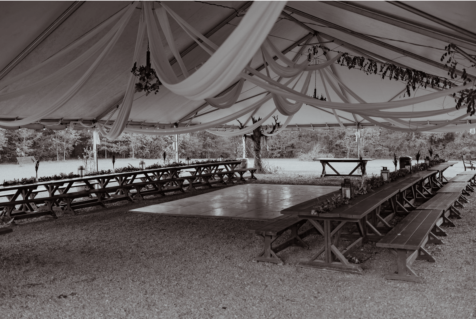 A large tent with draped fabric and string lights, surrounding a central dance floor with picnic tables on sides.