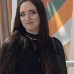 Young woman with long dark hair, wearing black clothing and layered necklaces, standing in front of colorful geometric background.