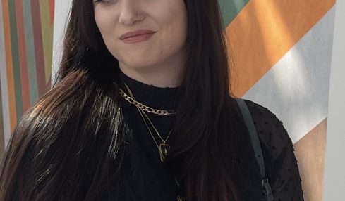 Young woman with long dark hair, wearing black clothing and layered necklaces, standing in front of colorful geometric background.