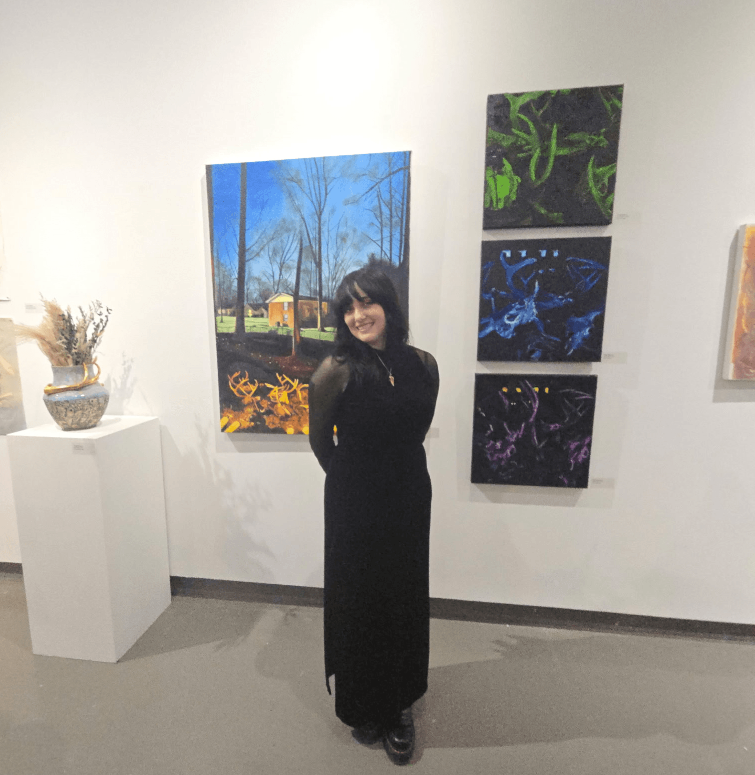 Woman in black dress standing in art gallery with colorful paintings on white wall behind her.