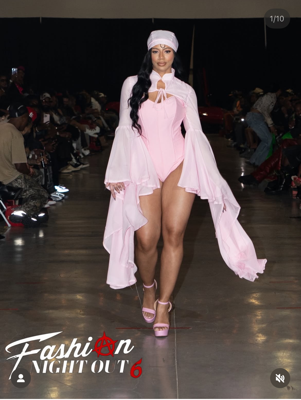Model walking on runway in pink outfit with flowing sleeves, headband, and high heels, audience seated on both sides.