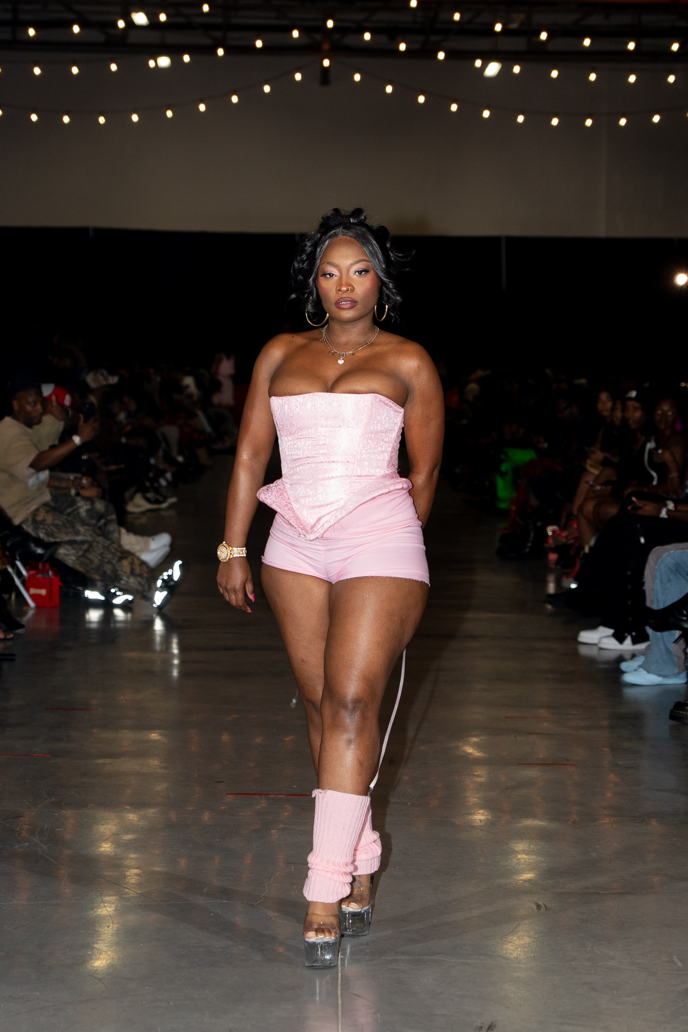 Woman walking on runway in pink strapless outfit with string lights overhead, audience seated on sides, polished floor.
