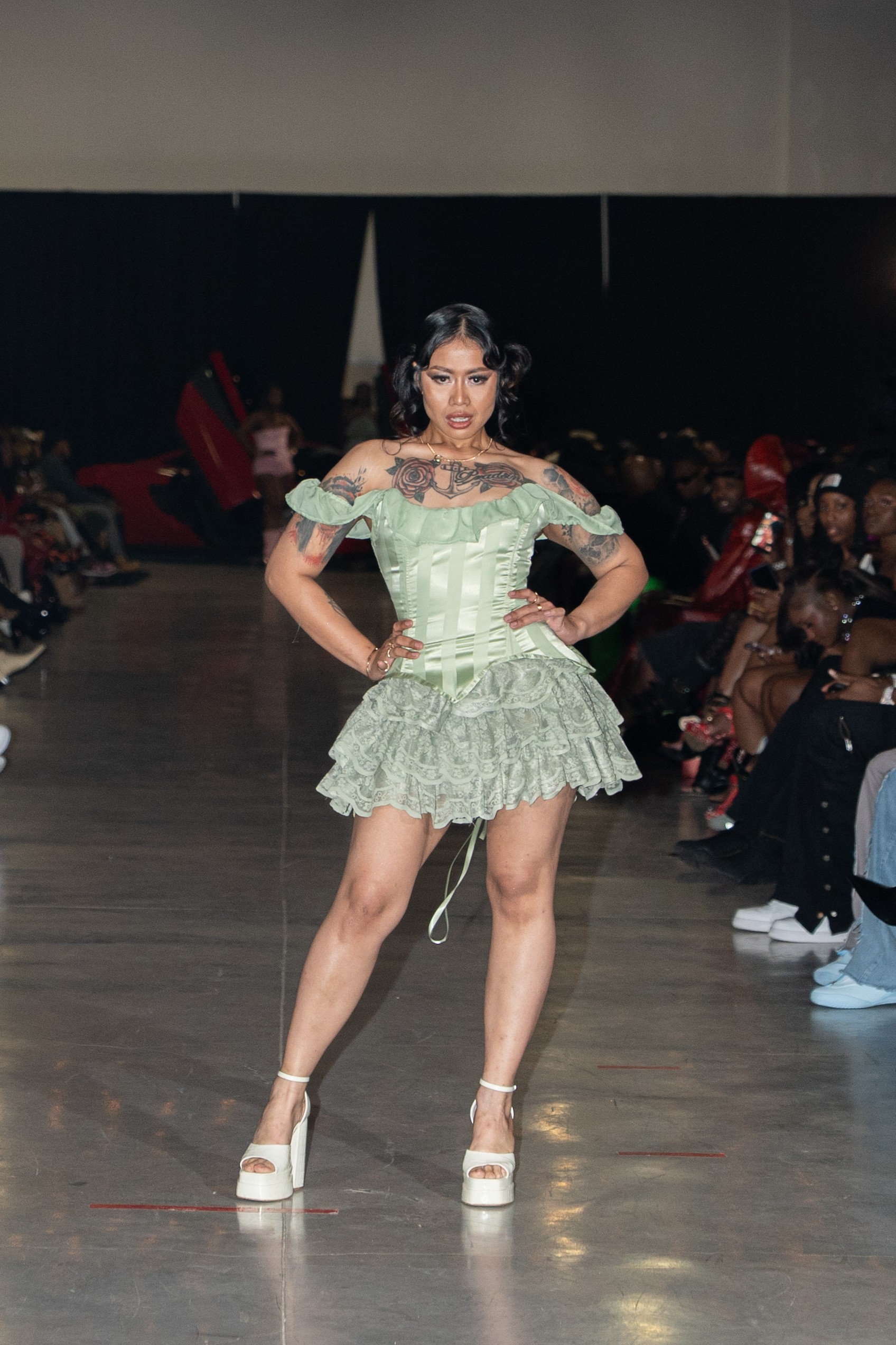 Model walking on runway in green ruffled dress with off-shoulder design, audience seated on both sides, dark background.