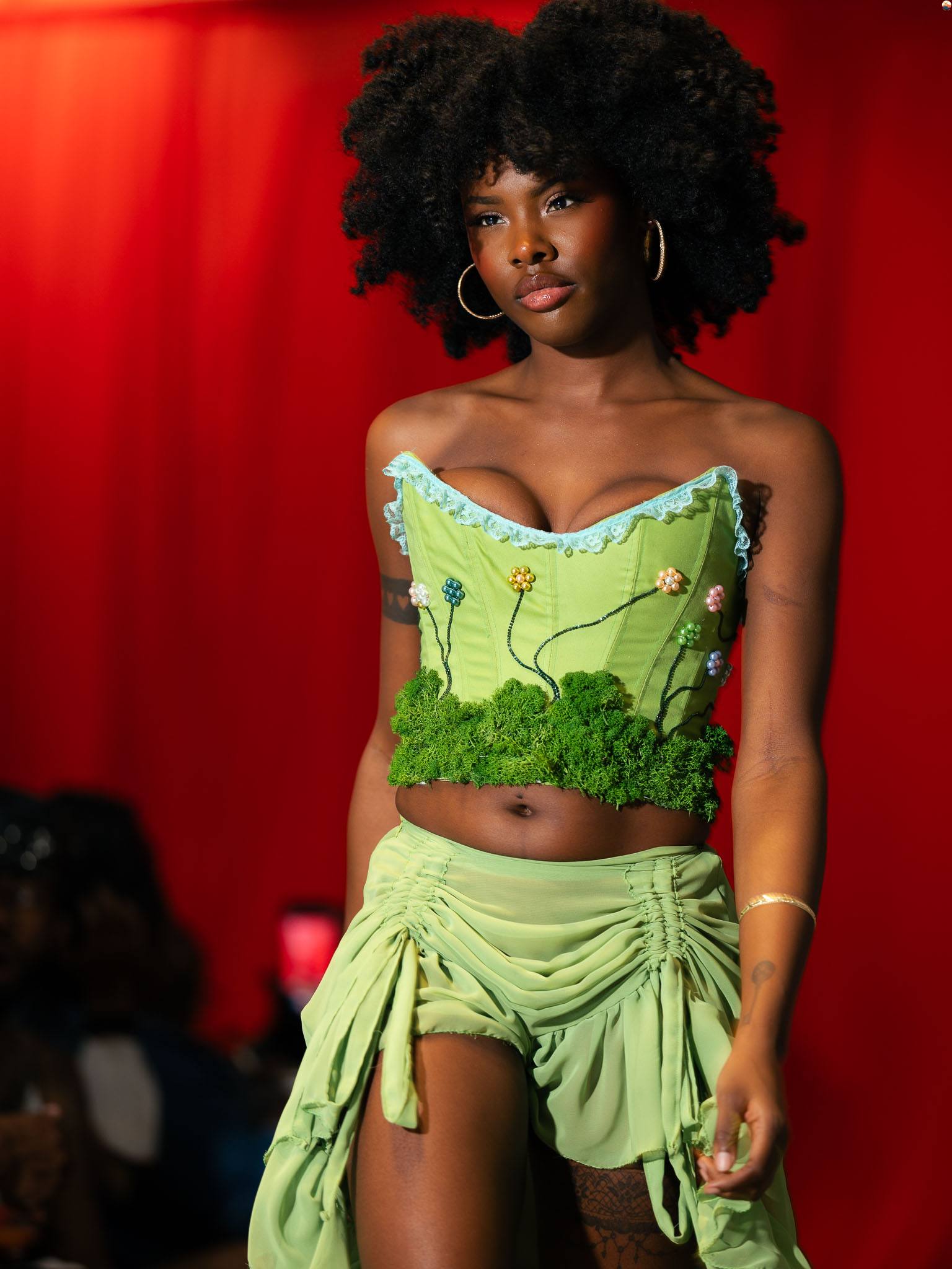 Young woman with natural curly hair wearing a green crop top and matching skirt on runway, red background.