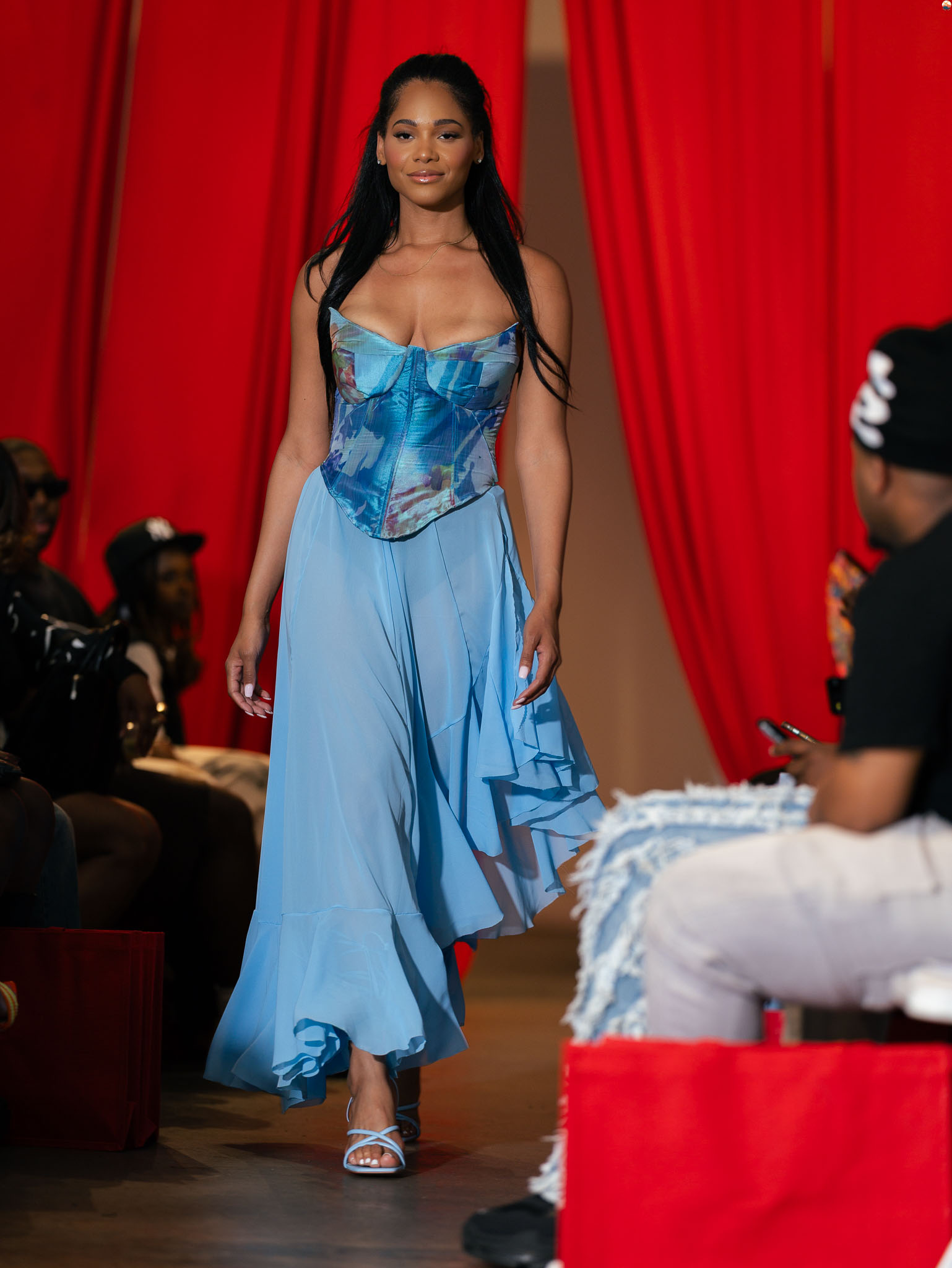 Woman walking on runway in blue dress with red curtains background, audience seated on sides, some taking photos.