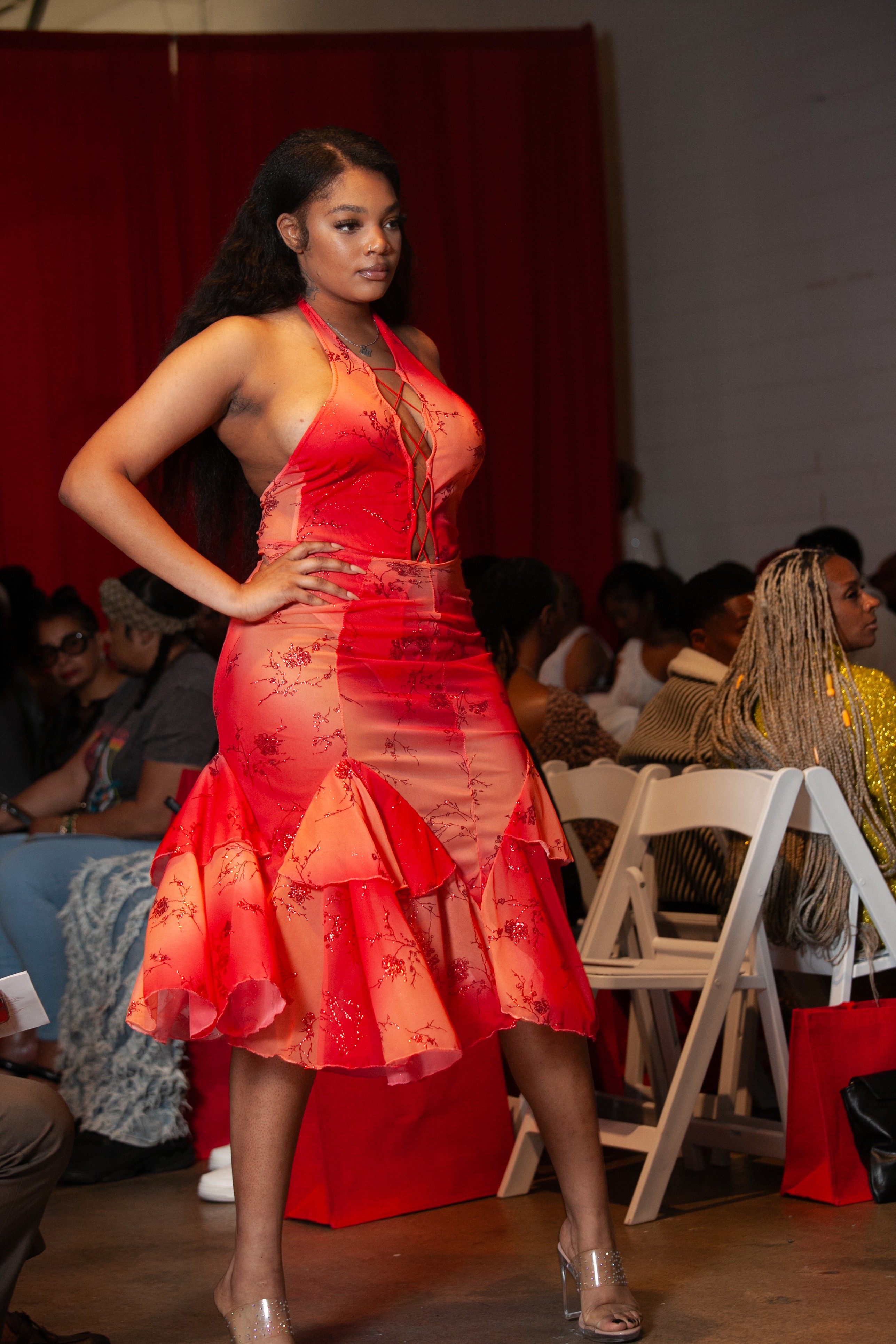 Woman in red dress walking on runway with audience in background, hands on hips, looking to the side.