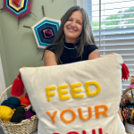 Smiling woman holding a pillow with yellow, orange, and red embroidered text, in a room with colorful wall decorations and a window.