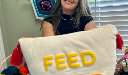 Smiling woman holding a pillow with yellow, orange, and red embroidered text, in a room with colorful wall decorations and a window.