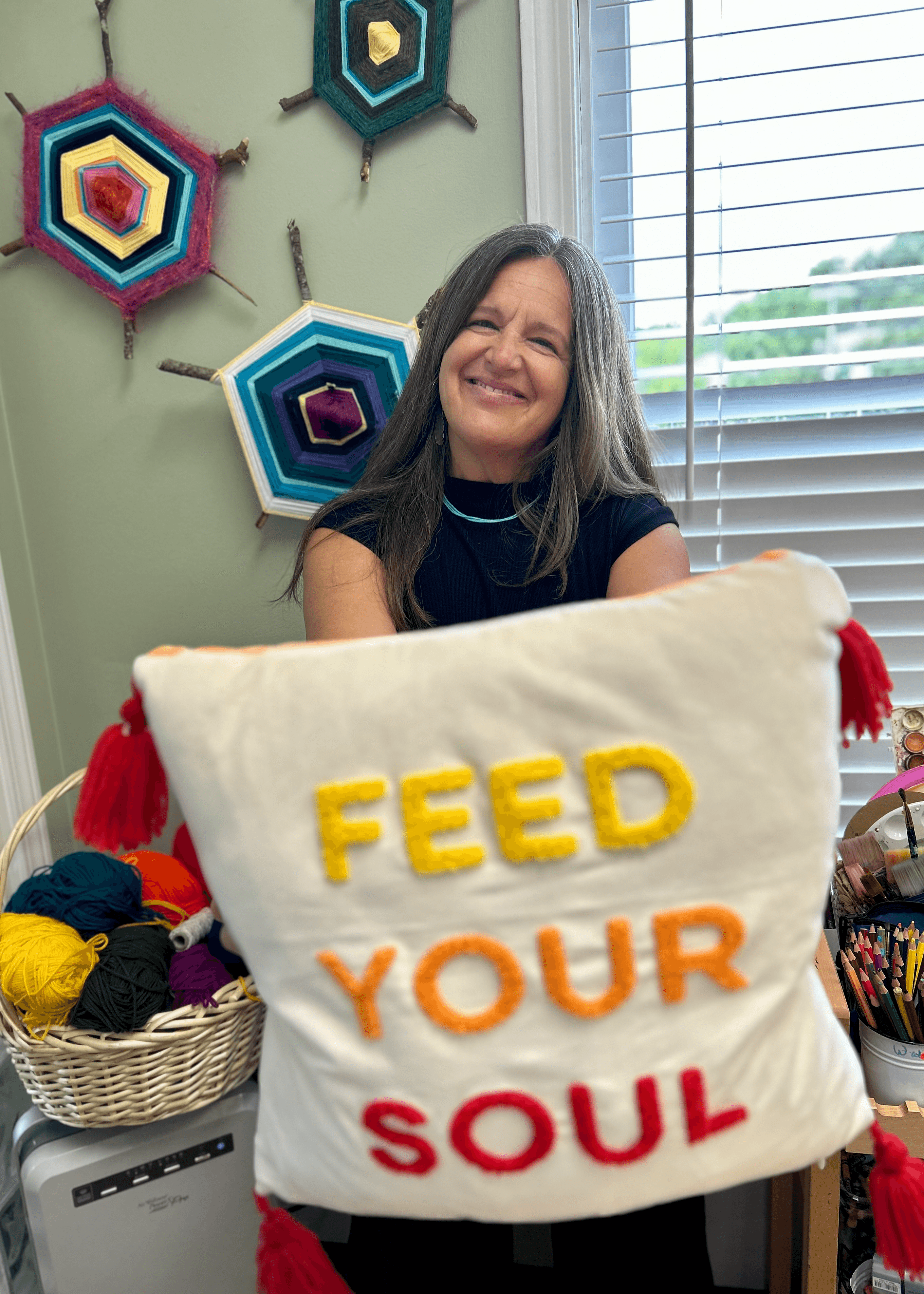 Smiling woman holding a pillow with yellow, orange, and red embroidered text, in a room with colorful wall decorations and a window.