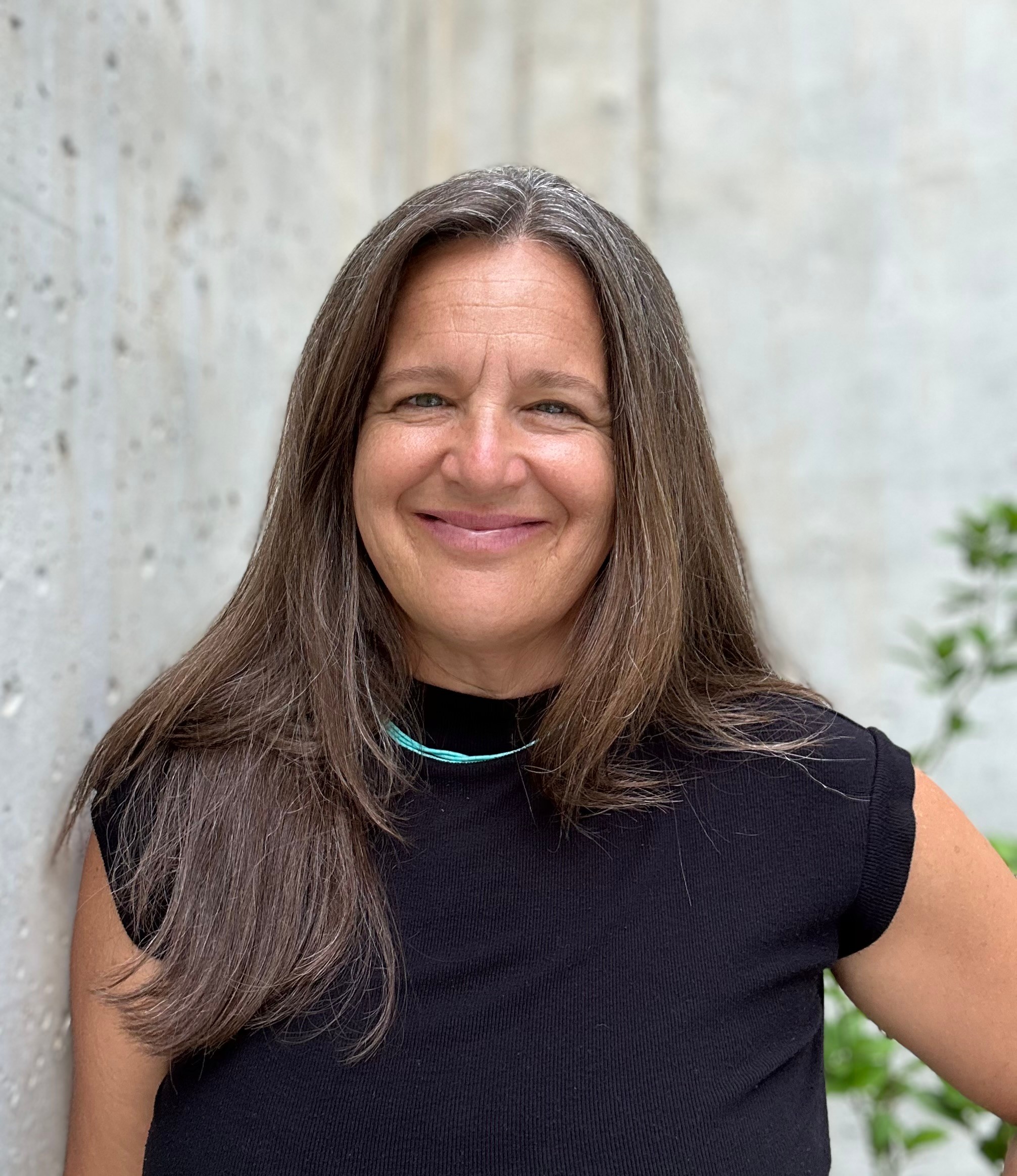 Smiling woman with long brown hair wearing a black top outdoors, with a concrete wall and greenery in the background.