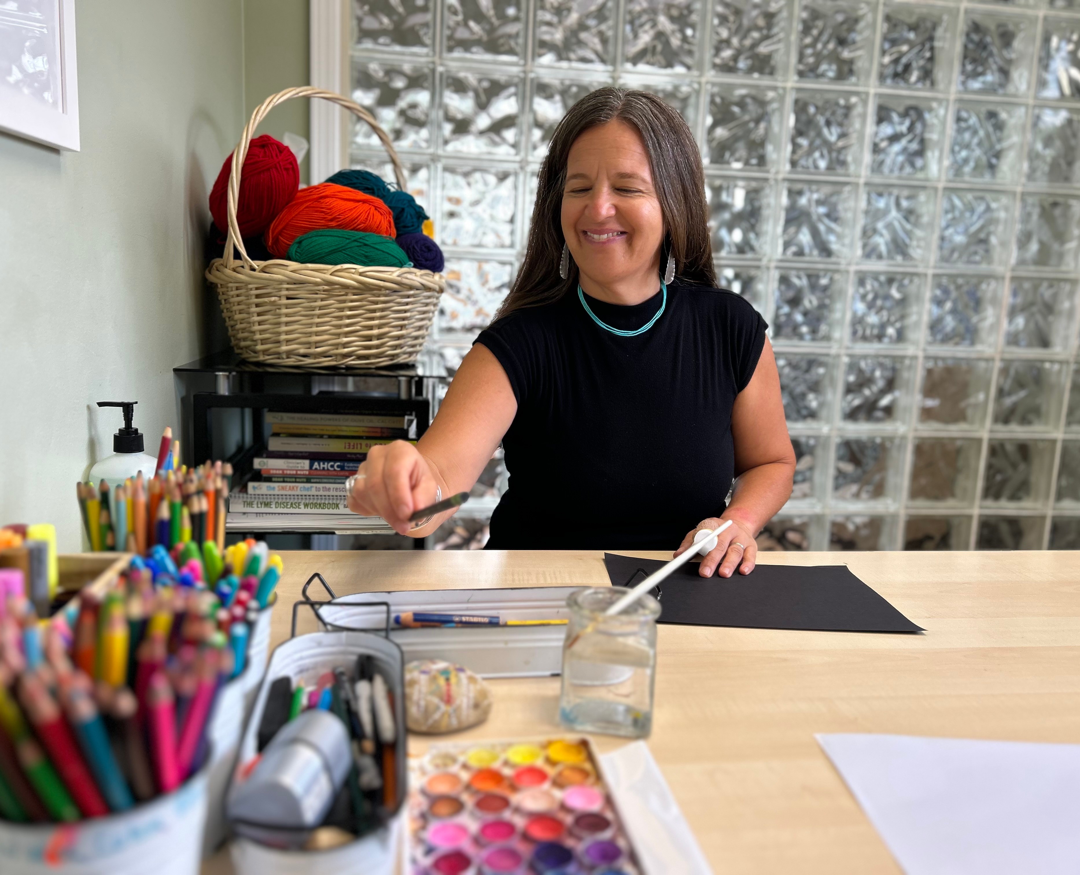 Woman smiling at a craft table with colorful supplies and a glass block, in a bright room with glass block wall.