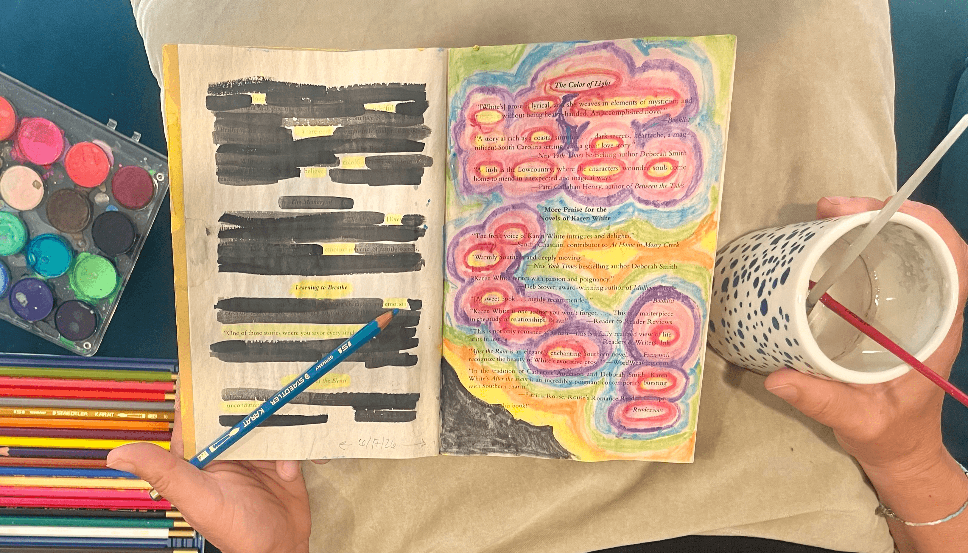 Open sketchbook with colorful drawings, surrounded by watercolor paints, colored pencils, and a cup with a paintbrush.