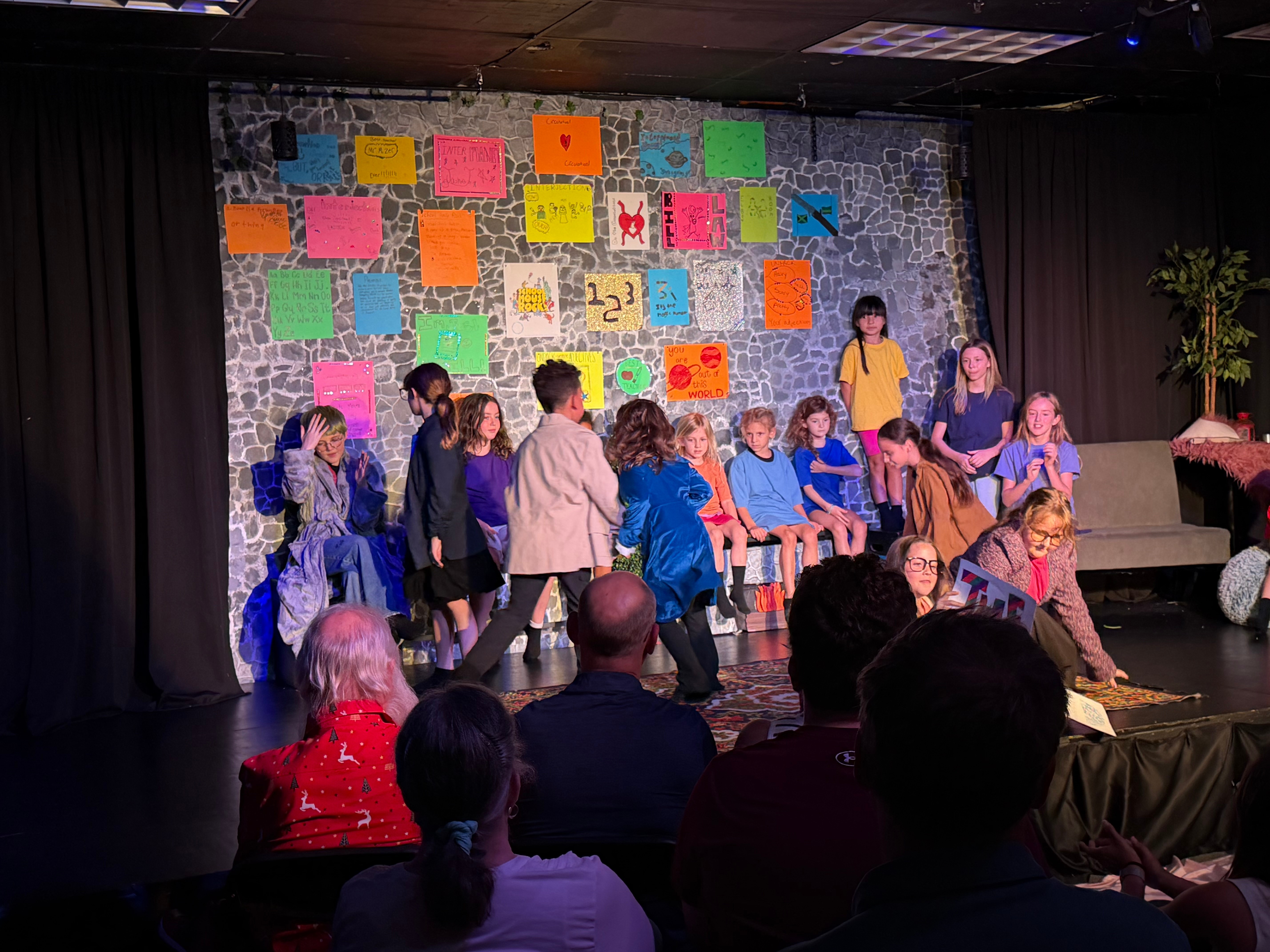 Children perform on stage with colorful posters on the wall, audience watching, some children seated, one standing, in a theater setting.