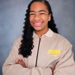 Young girl with curly hair smiling, arms crossed, wearing a beige jacket with yellow text, against a gray background.