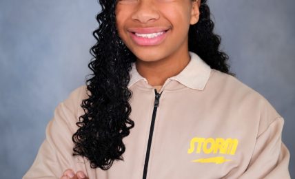 Young girl with curly hair smiling, arms crossed, wearing a beige jacket with yellow text, against a gray background.