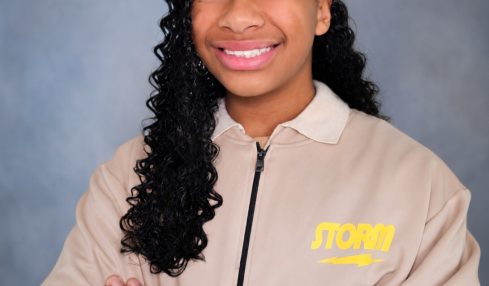 Young girl with curly hair smiling, arms crossed, wearing a beige jacket with yellow text, against a gray background.