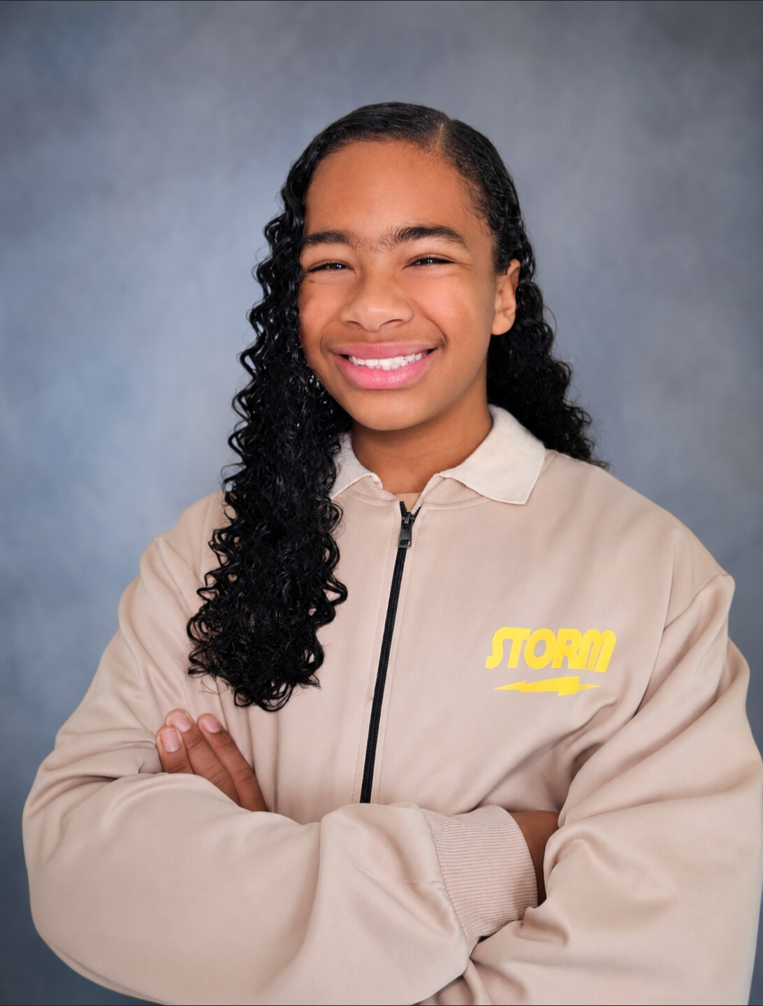 Young girl with curly hair smiling, arms crossed, wearing a beige jacket with yellow text, against a gray background.