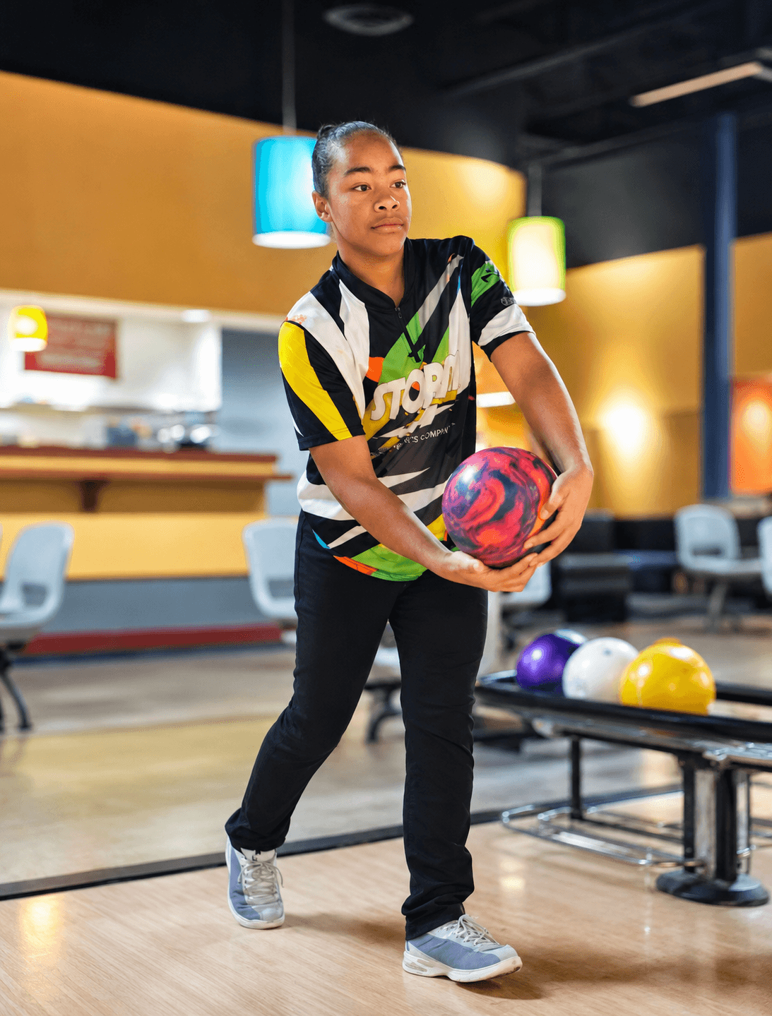 Young woman holding a colorful bowling ball in a bowling alley with bowling balls on a rack behind her.