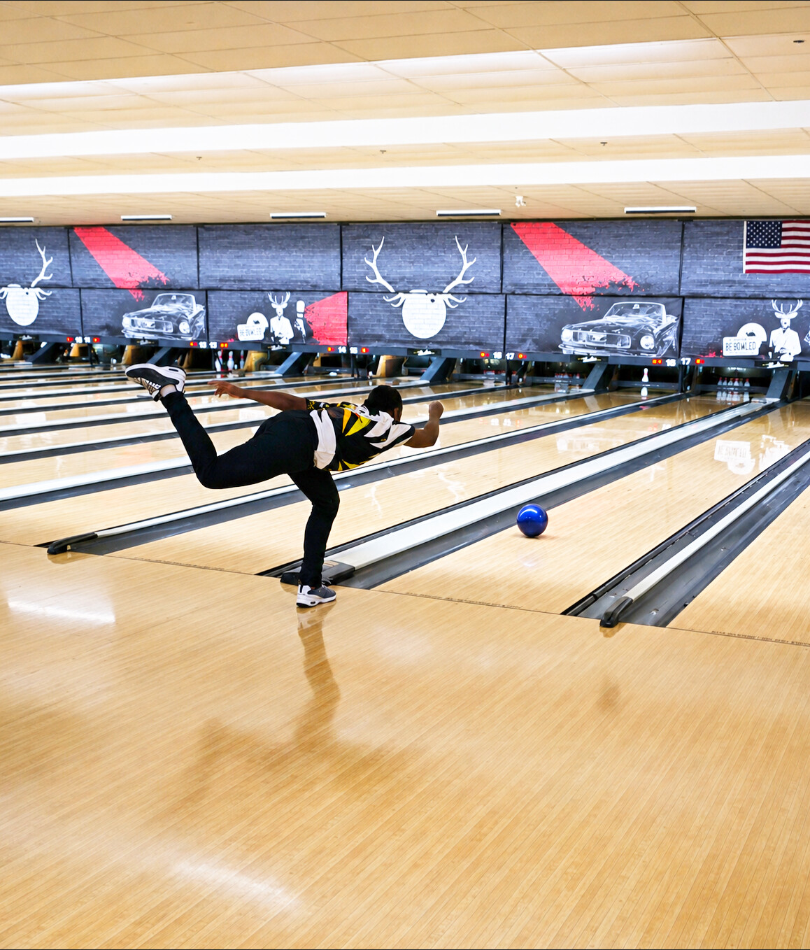 Person bowling on a lane, releasing a ball, with a decorated wall and American flag in the background.