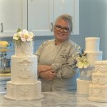 Smiling woman with glasses and gray hair in a kitchen, surrounded by white decorated cakes on a marble counter.