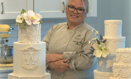 Smiling woman with glasses and gray hair in a kitchen, surrounded by white decorated cakes on a marble counter.