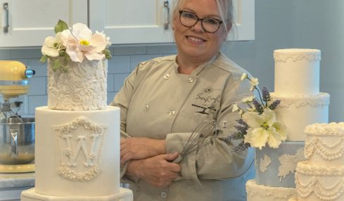 Smiling woman with glasses and gray hair in a kitchen, surrounded by white decorated cakes on a marble counter.