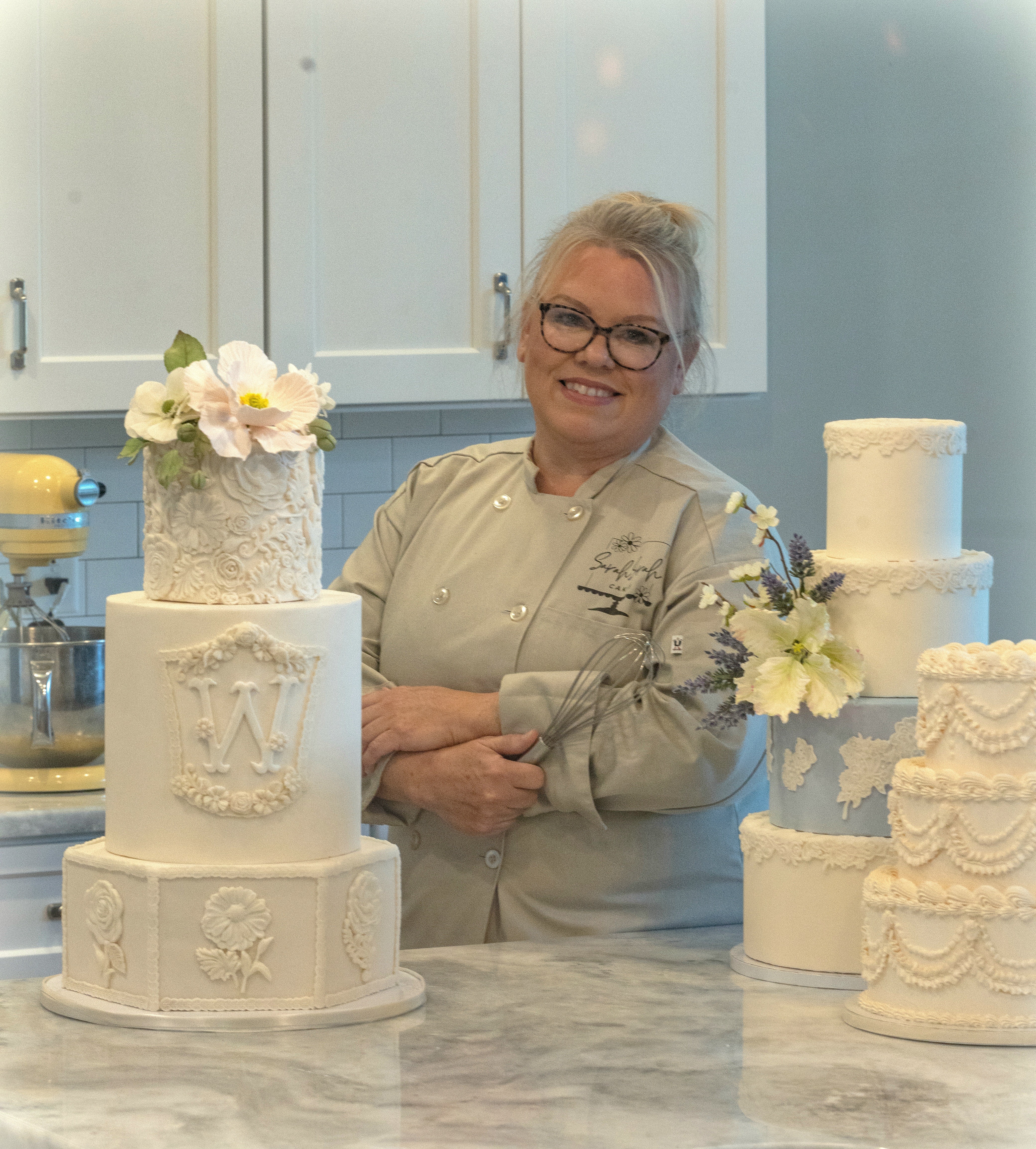 Smiling woman with glasses and gray hair in a kitchen, surrounded by white decorated cakes on a marble counter.