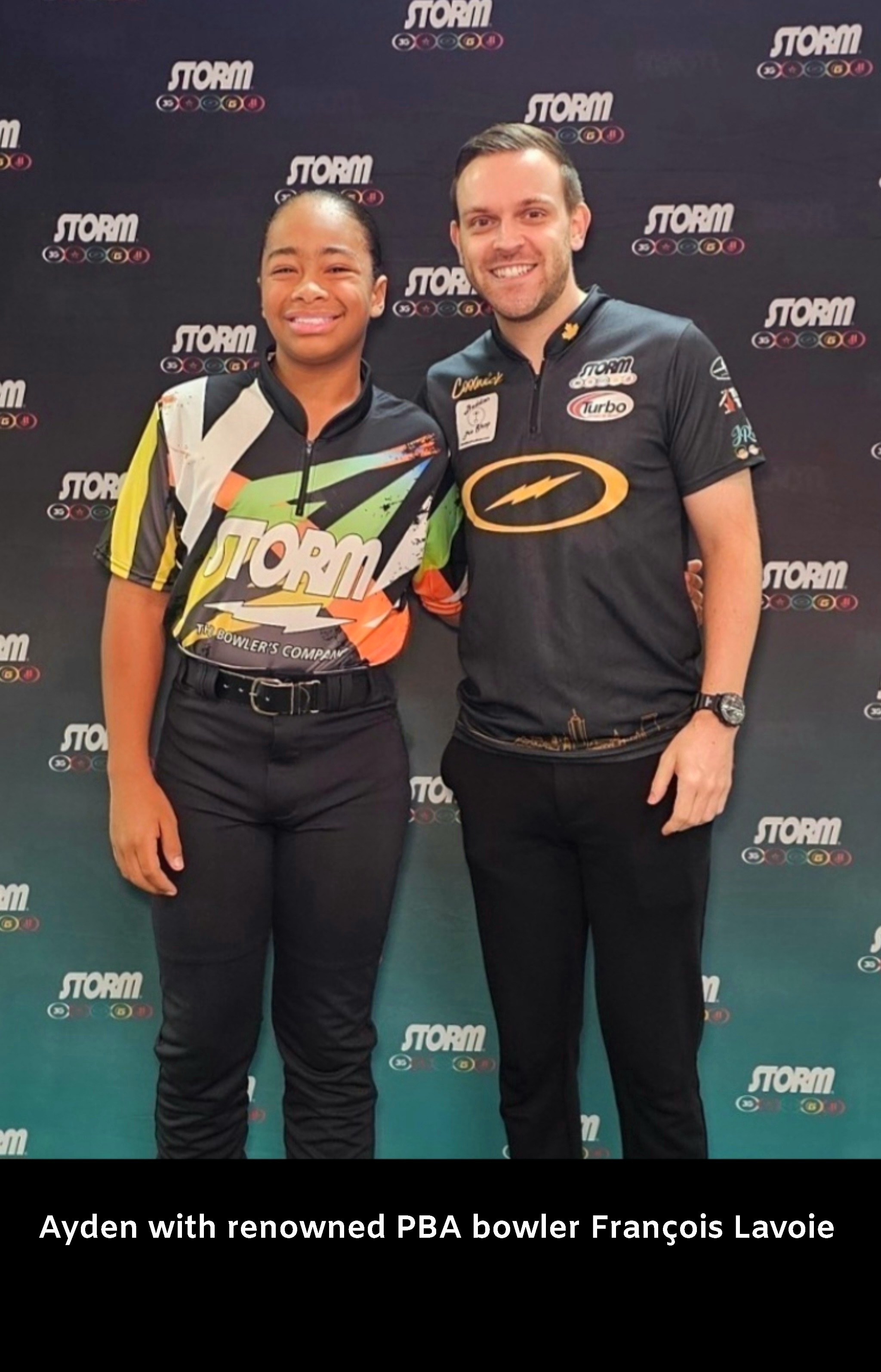 Two people, a woman and a man, stand together smiling in front of a backdrop with logos. Woman wears a colorful shirt, man wears a black sports shirt.
